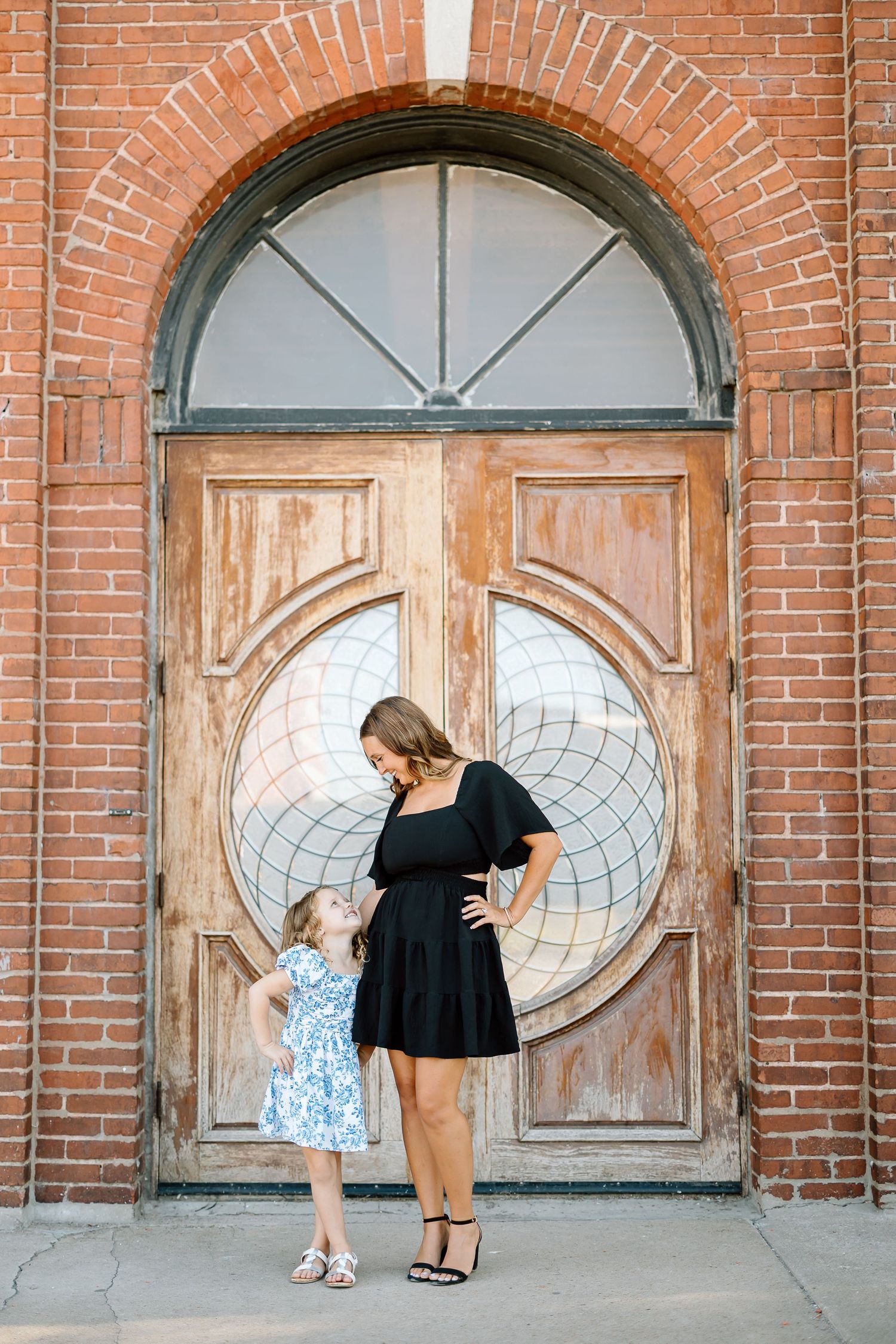 Two people stand in front of a grand wooden door with circular window detail and brick archway entrance.
