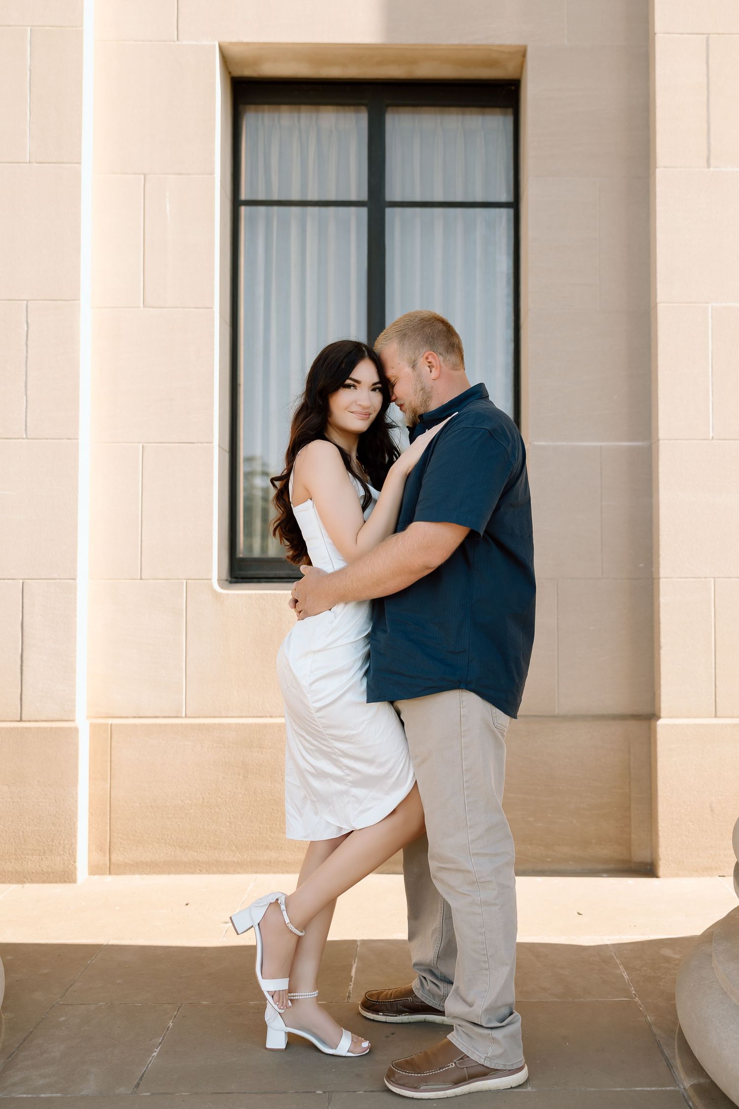 A romantic couple embraces near a tall window, with the woman wearing a white dress and the man in a navy shirt.
