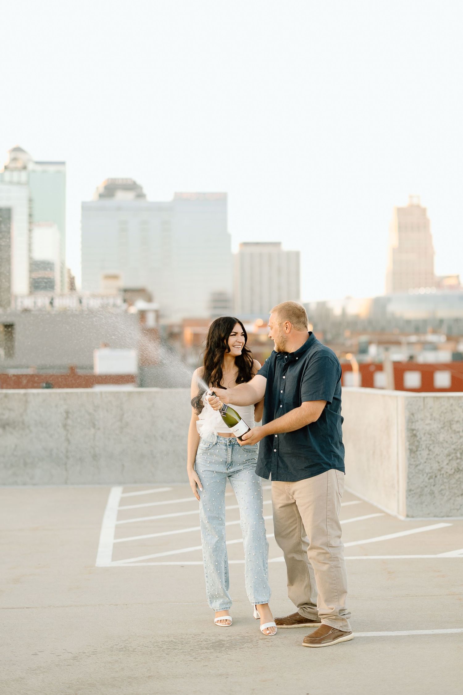 A couple shares a romantic moment on a rooftop parking deck with a city skyline in the background at sunset.