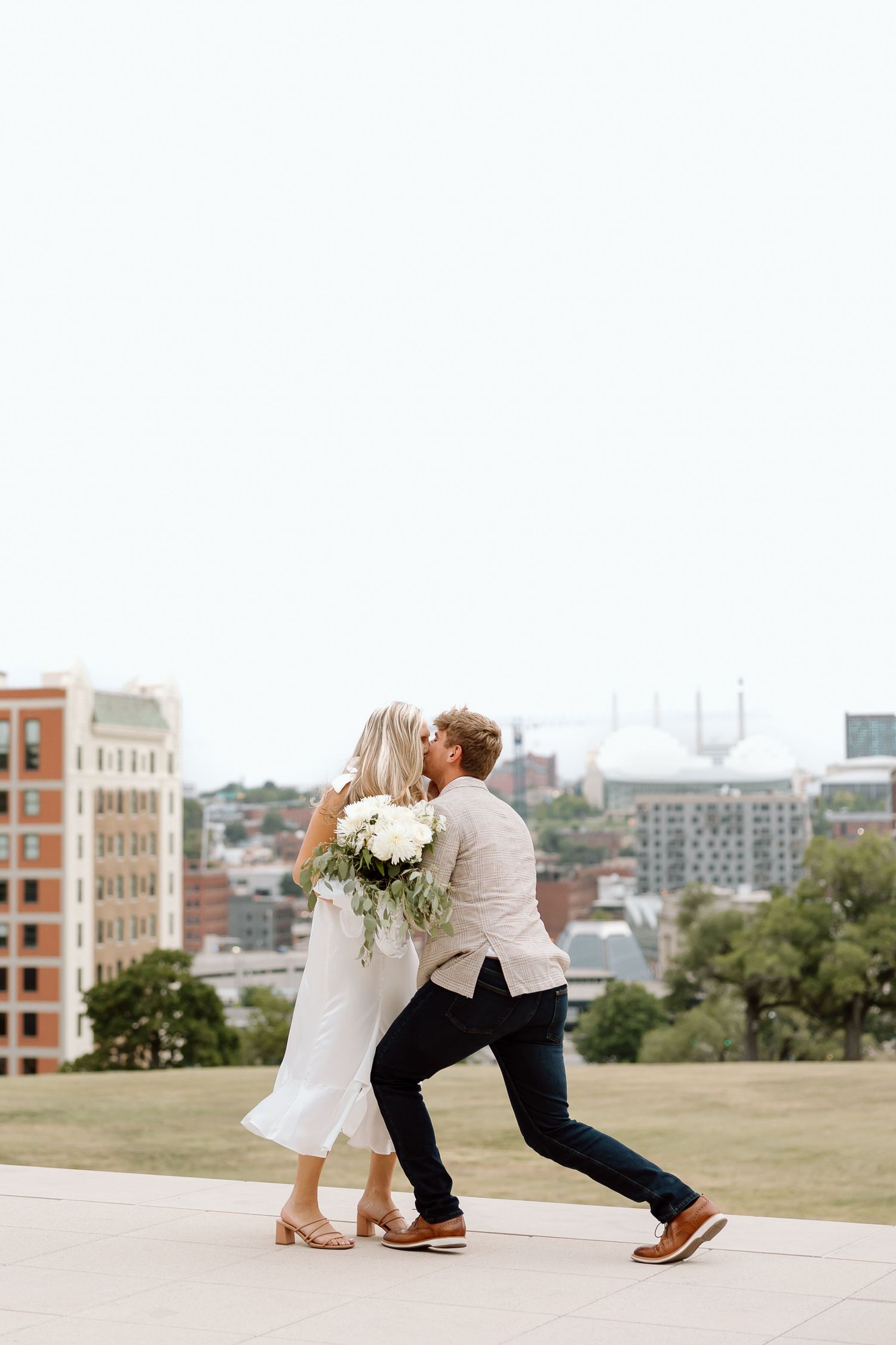 A couple dips and embraces during their romantic photoshoot on a grassy hill overlooking a cityscape.