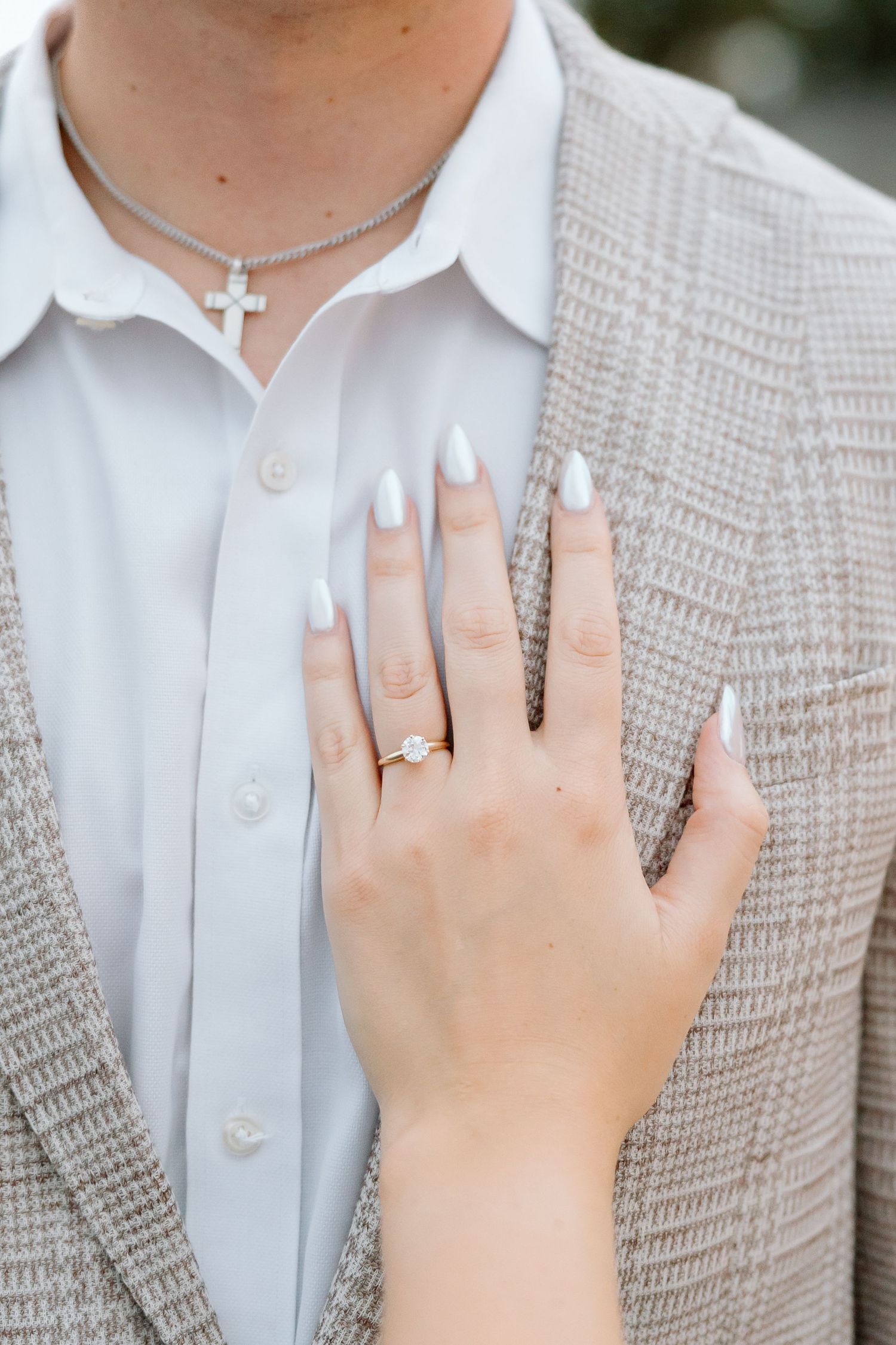 Close-up of a hand with white manicured nails wearing an engagement ring against a white shirt and beige cardigan.