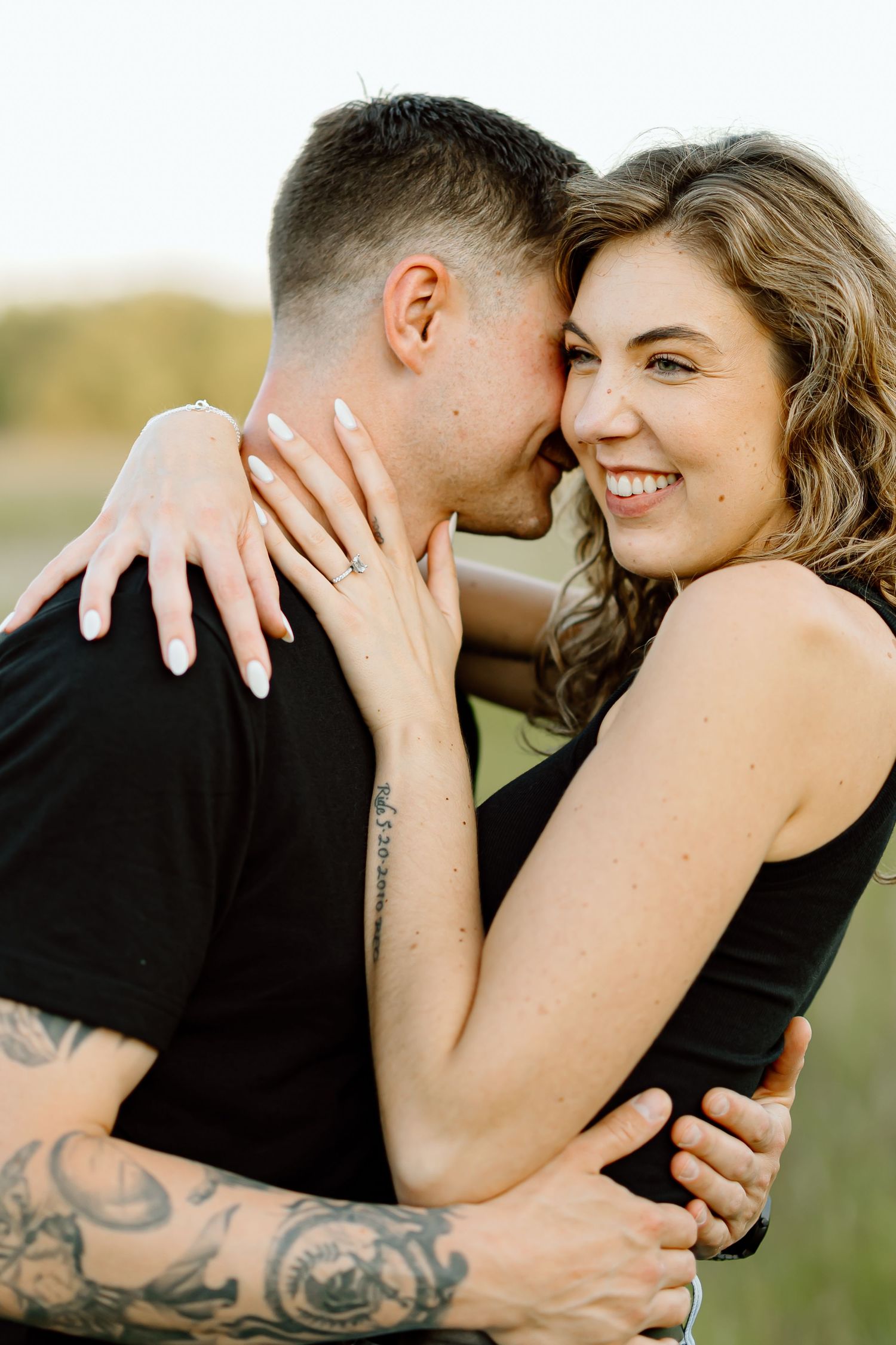 A romantic couple embraces outdoors during a sunset photoshoot, with visible tattoo details.