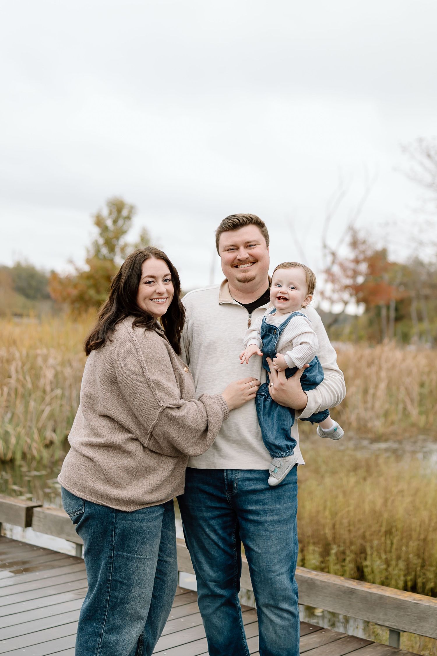 A family stands together on a wooden boardwalk surrounded by autumn grasses on an overcast day.