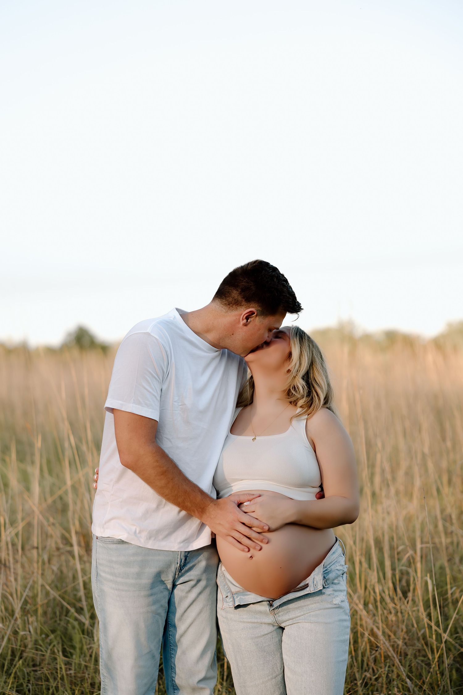 A couple embraces tenderly in a golden wheat field during sunset, highlighting the woman's pregnant belly.