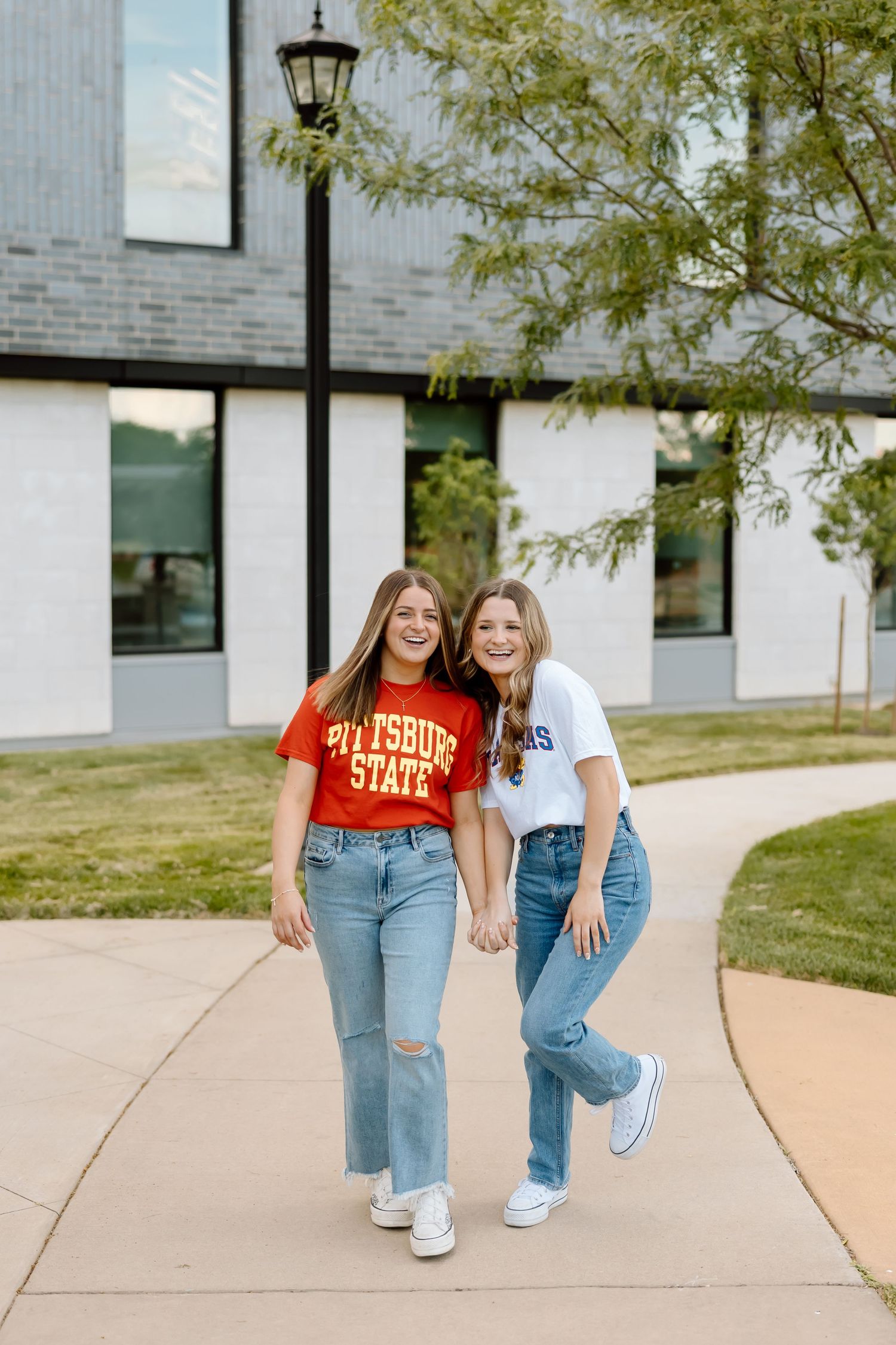 Two friends in casual outfits posing together on a concrete sidewalk with white buildings and trees in background.