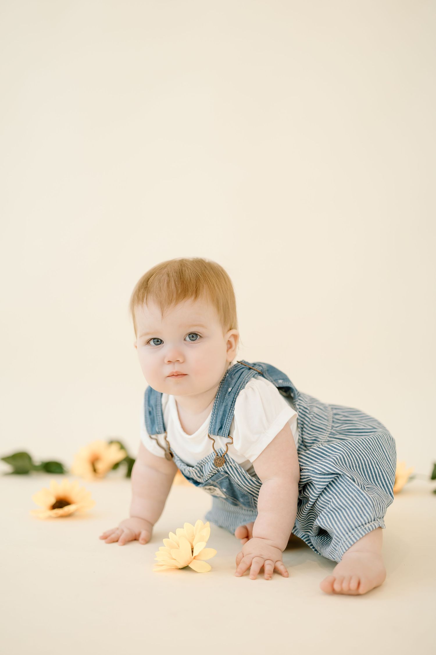 A baby in striped overalls crawls on a light colored backdrop with soft peach flowers in the background.