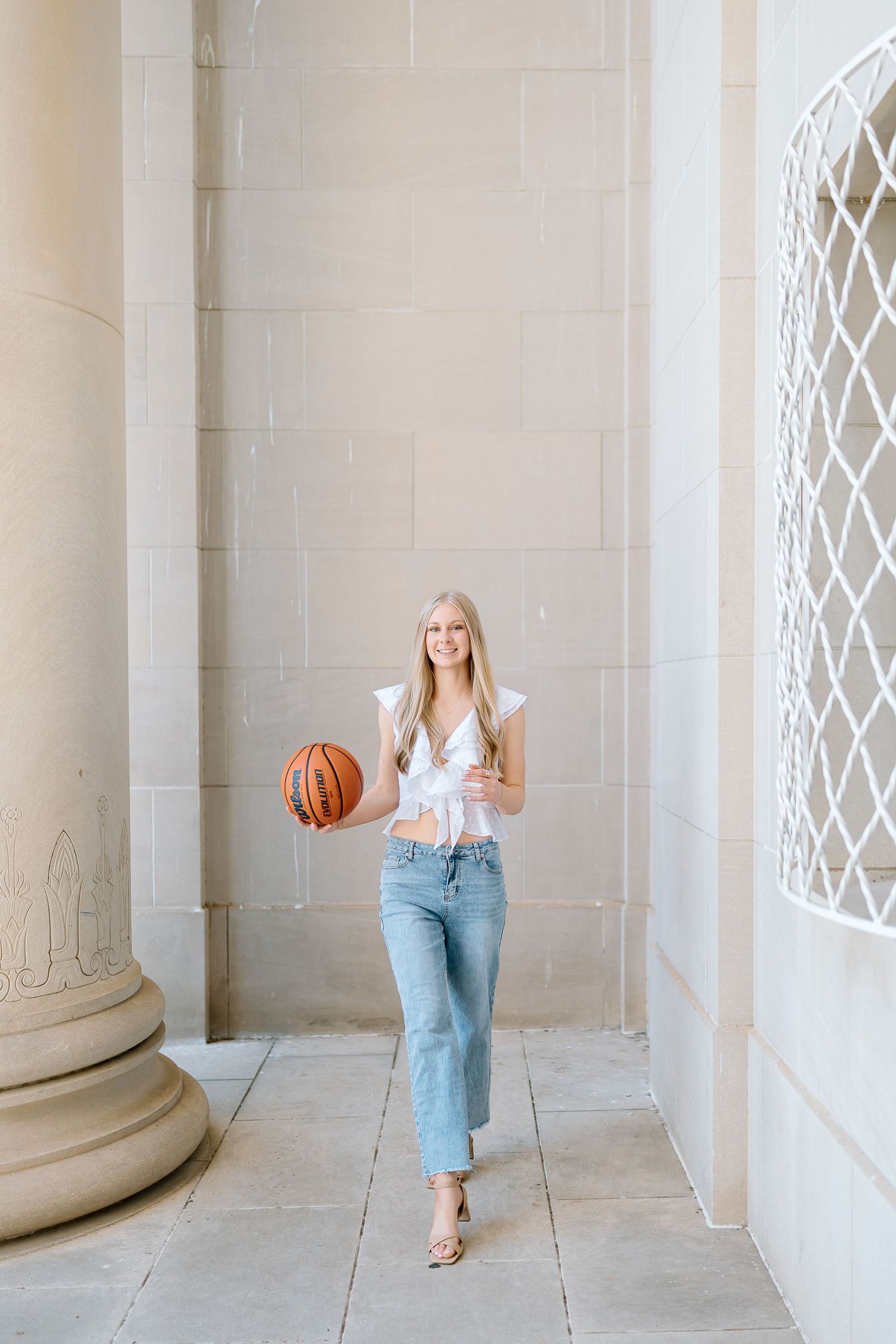 A person in casual denim and white top holds a basketball while walking through an elegant columned hallway.