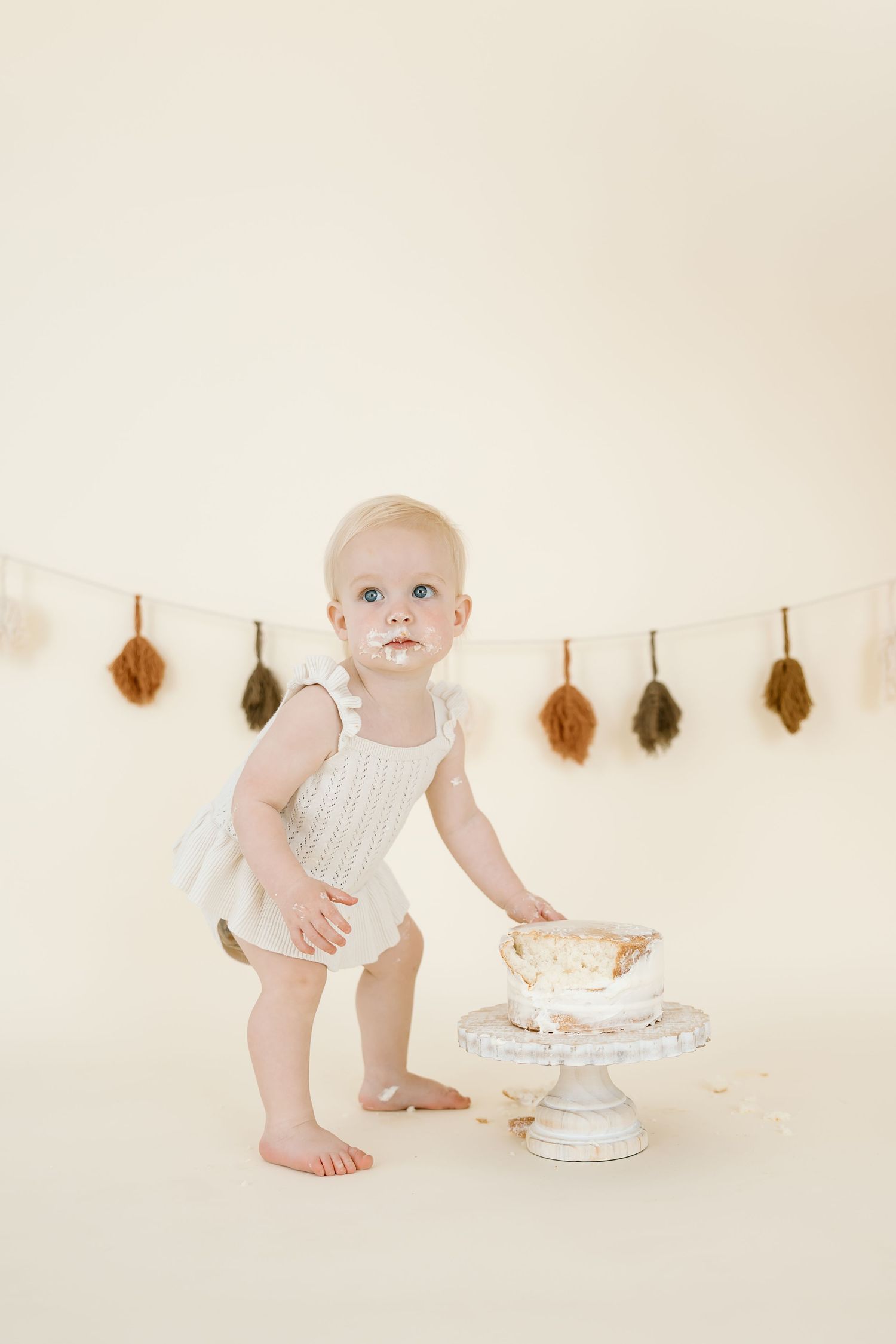 Baby standing beside white cake with dried leaf decorations hanging on neutral wall.