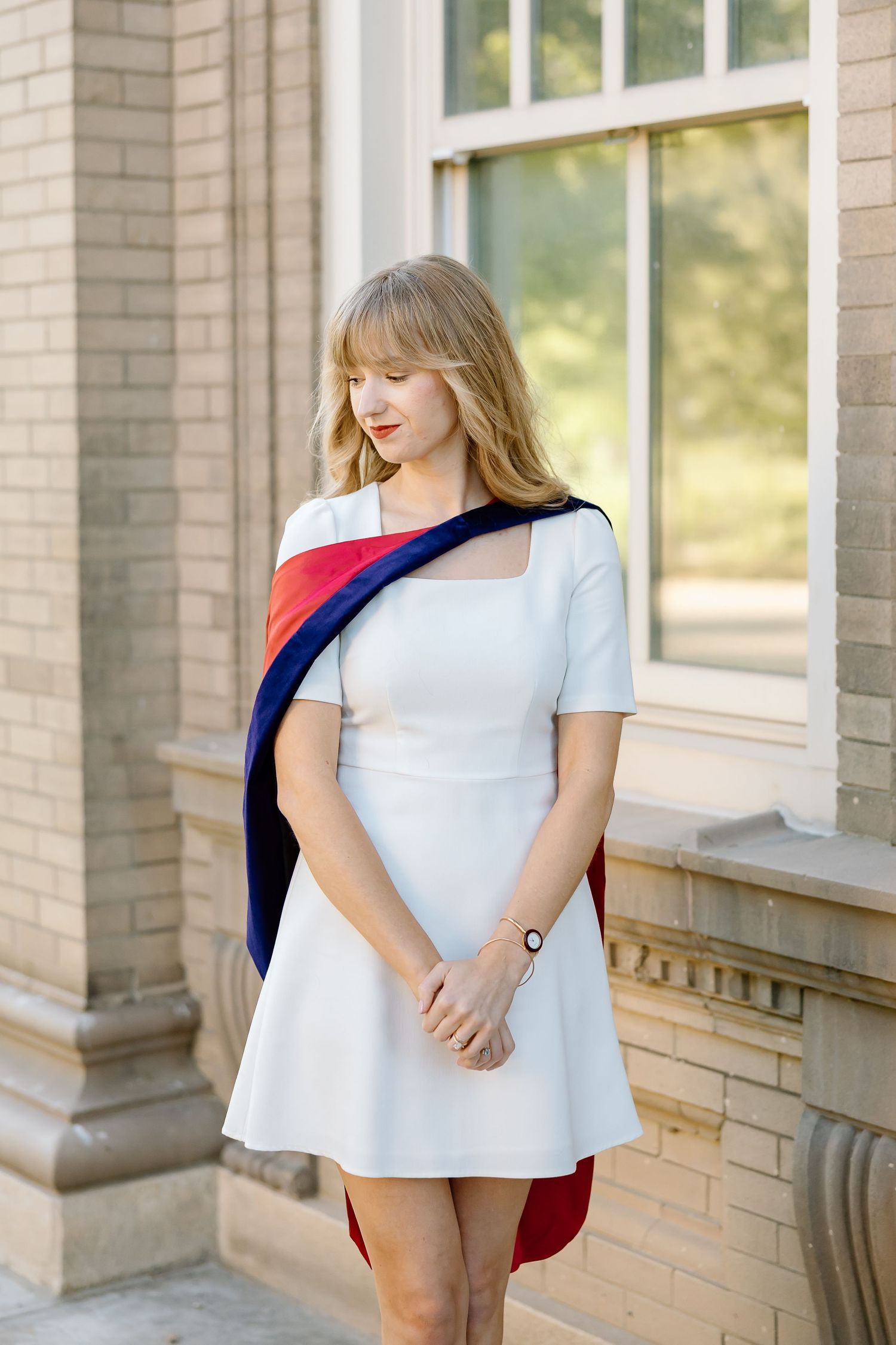A woman poses in an elegant white dress with a navy and red accent piece against a brick building exterior.