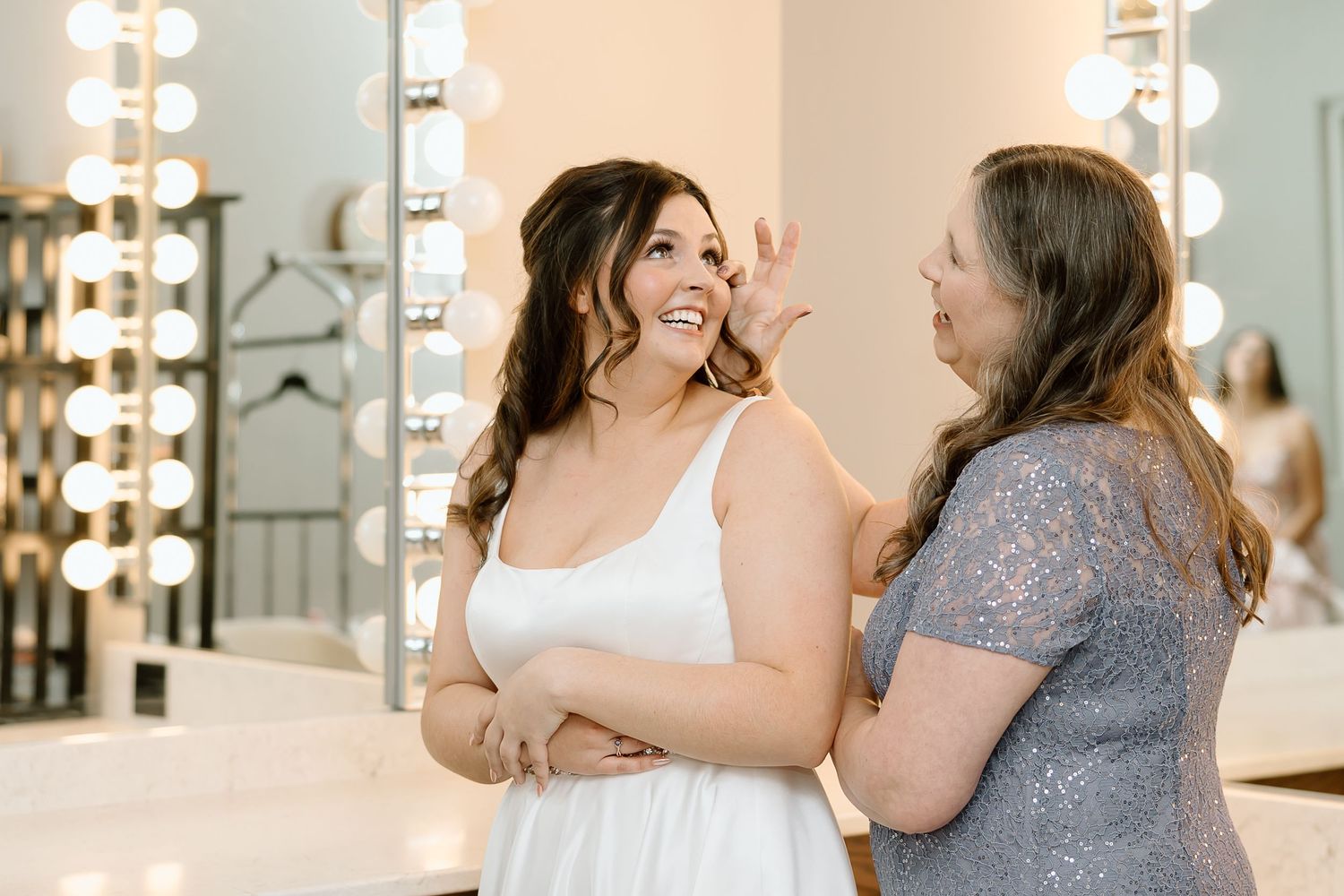 Two women in white and gray dresses share a joyful moment in front of illuminated vanity mirrors.