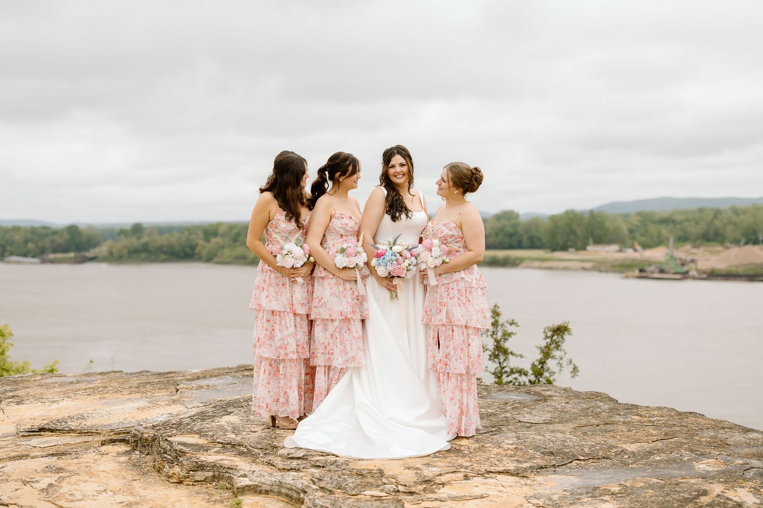 A wedding party in blush pink dresses stands together on a scenic rocky beach overlook at sunset.