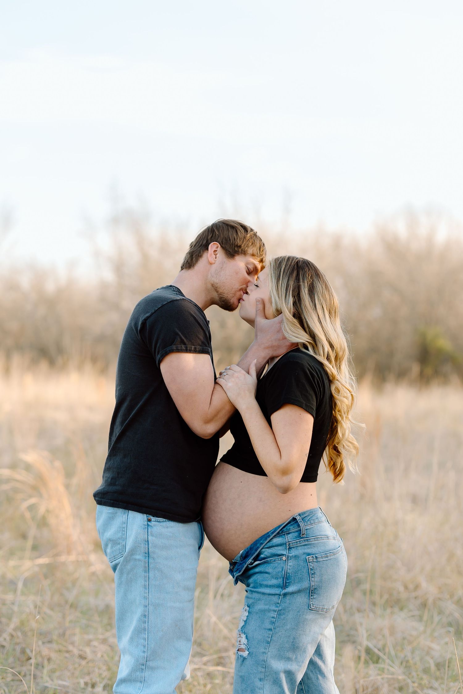 A couple in matching black t-shirts and jeans embrace lovingly in a sunlit field at golden hour.