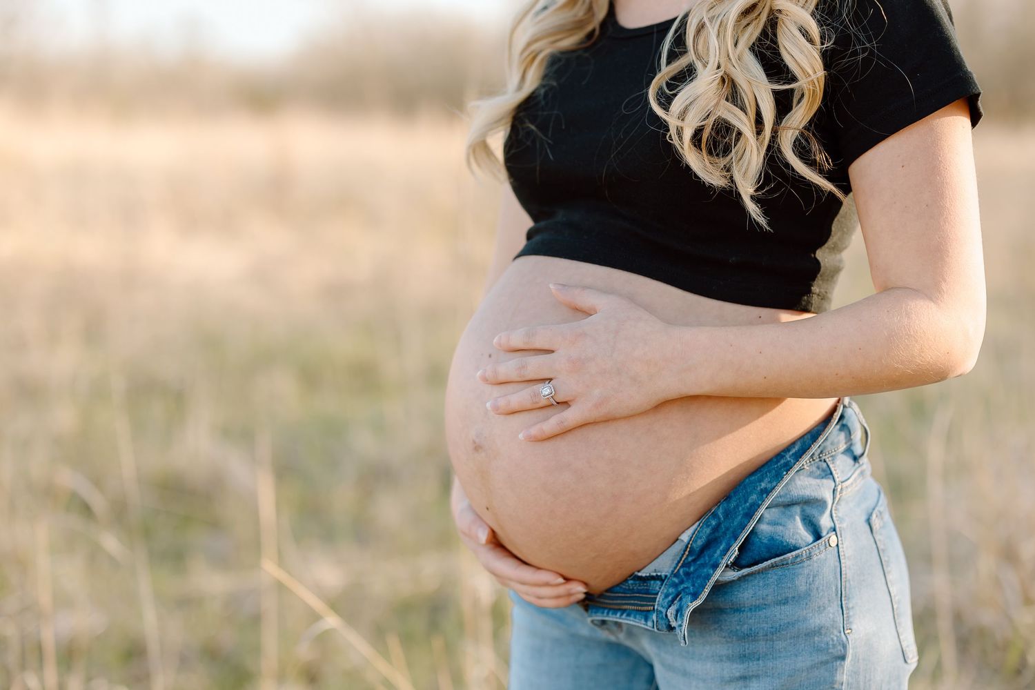 A pregnant woman in a black top and jeans cradles her baby bump while standing in a sunlit field at golden hour.