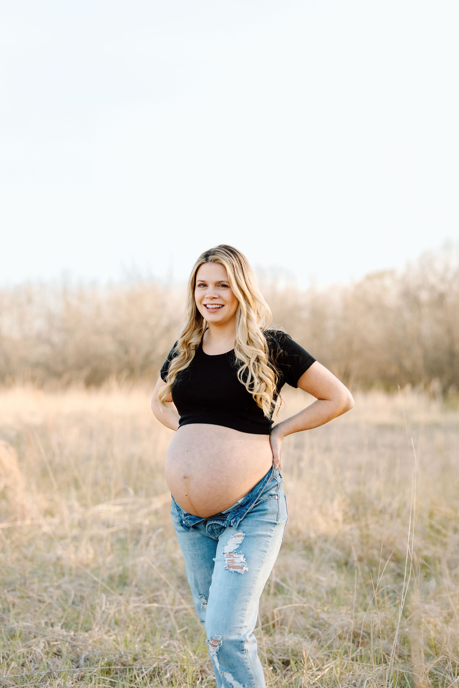 A maternity photoshoot in a golden field shows someone wearing a black top and distressed jeans at sunset.