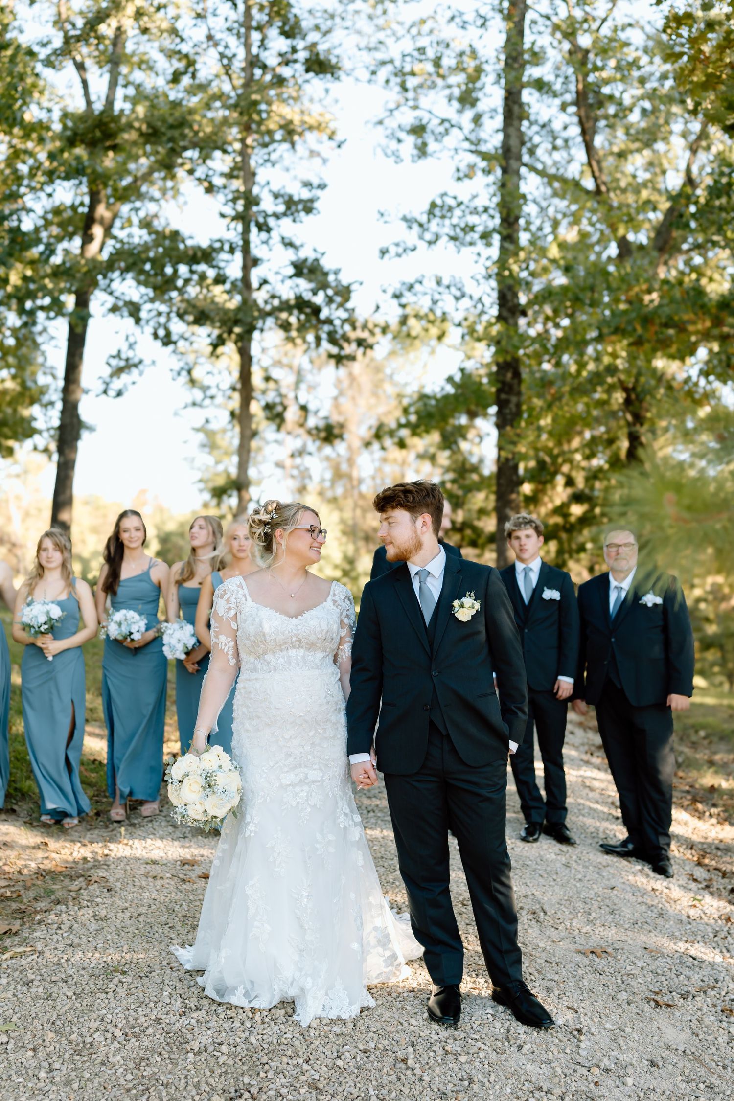 Wedding party walks along gravel path with bridesmaids in teal dresses and groomsmen in black suits during outdoor ceremony.