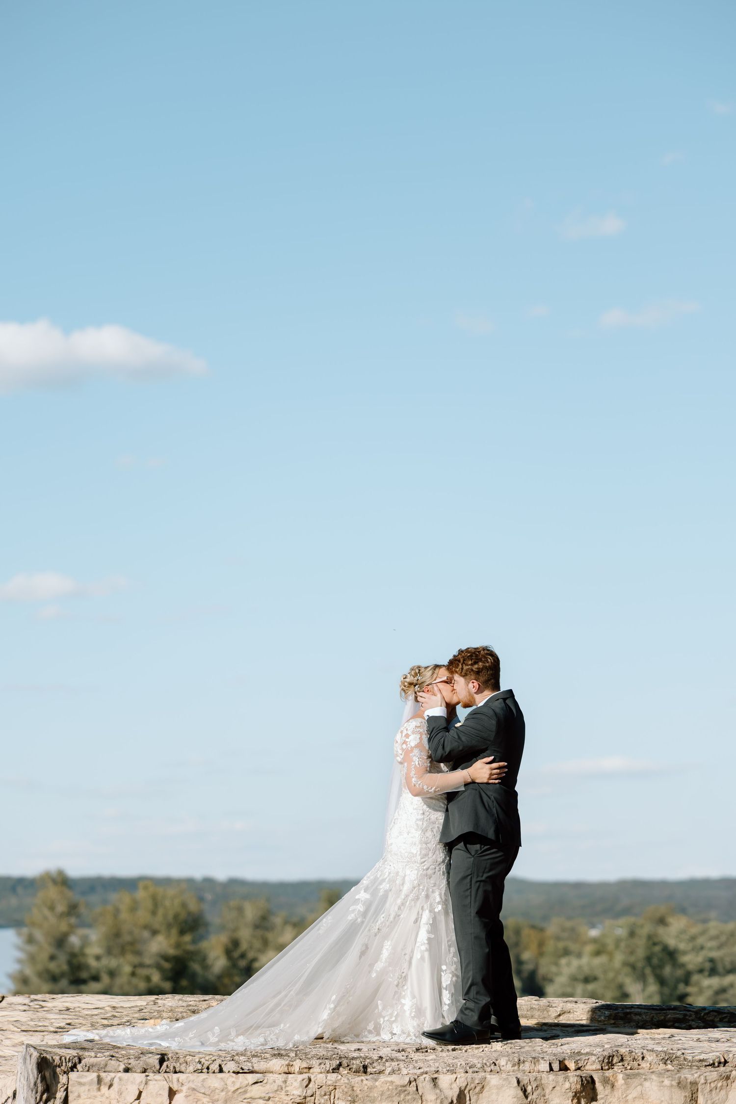 A romantic wedding moment captured as newlyweds embrace on a stone overlook with rolling hills in the background.
