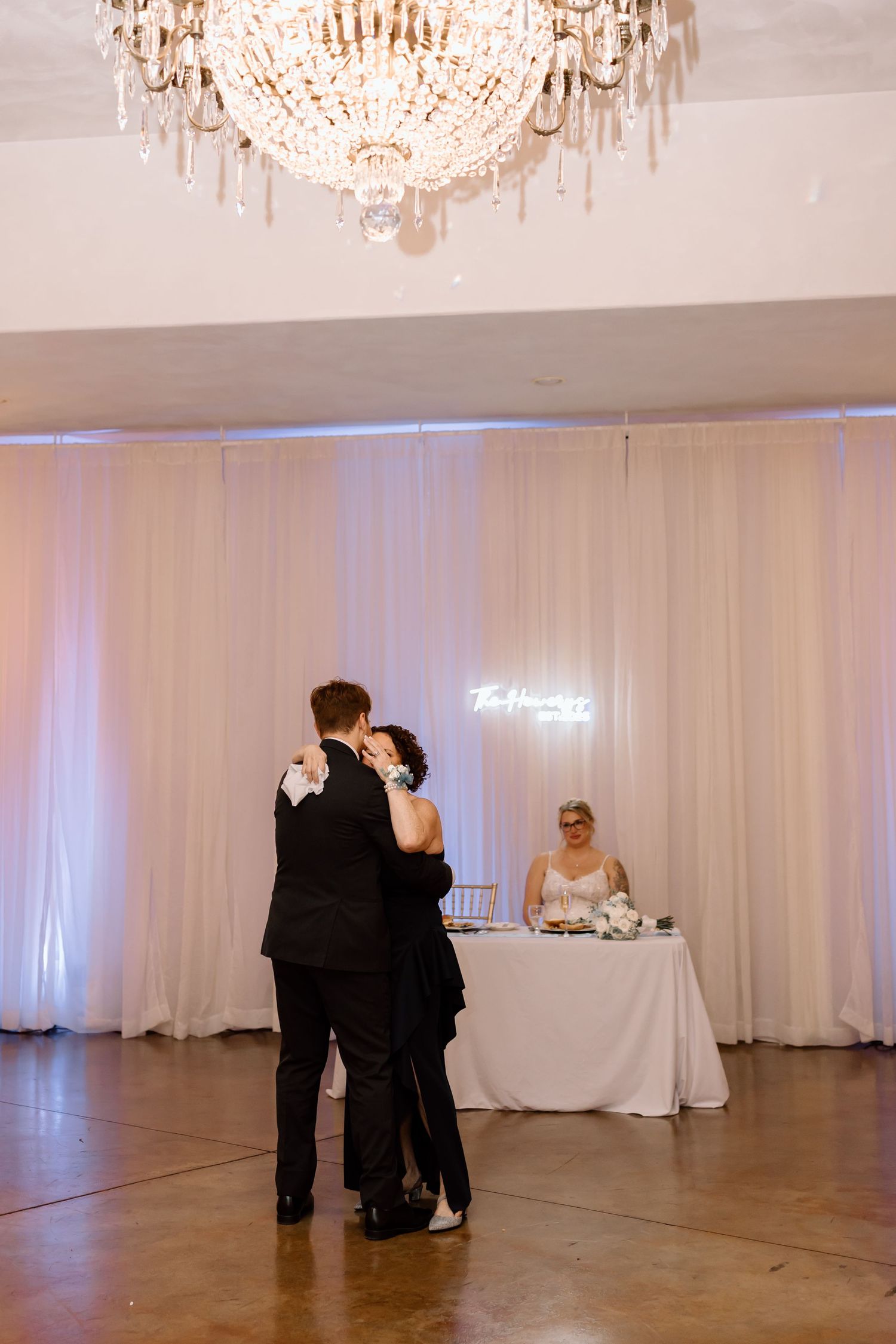 Wedding guests dance in an elegant ballroom with white drapes and crystal chandeliers overhead.