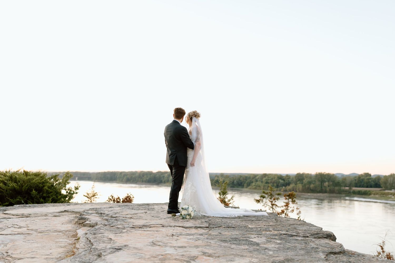 A couple in wedding attire stands on a rocky shoreline overlooking a serene lake during their outdoor celebration.