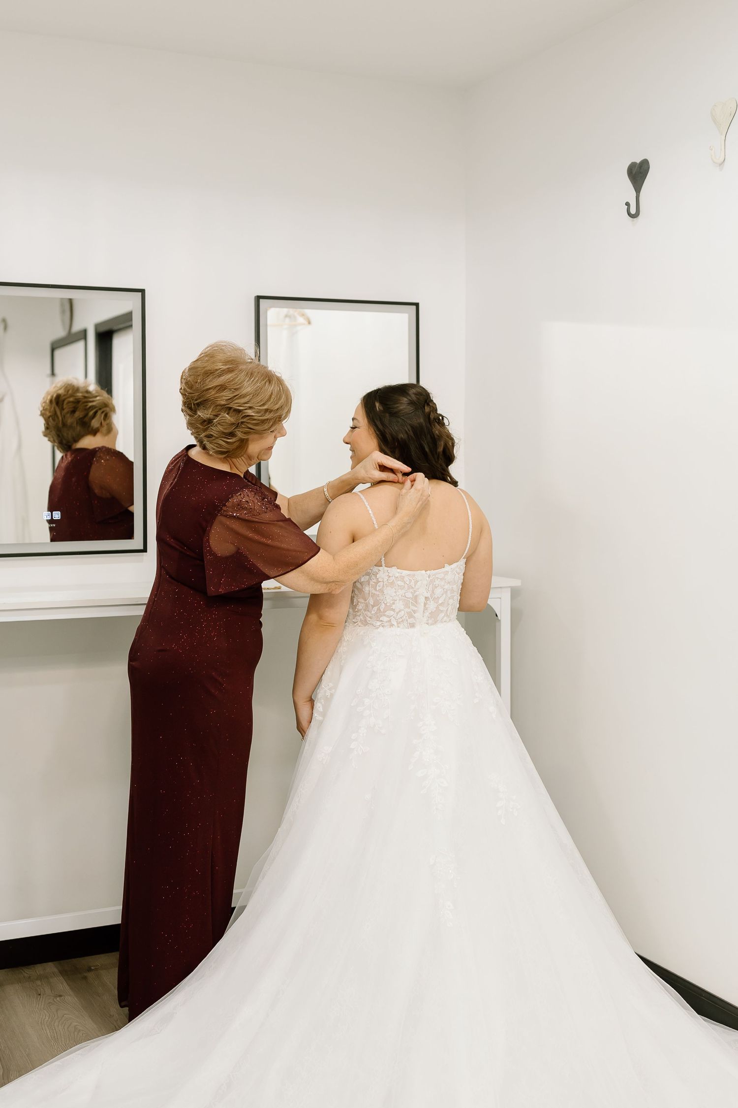 A woman in a white wedding dress gets help with final adjustments in a bright white room.