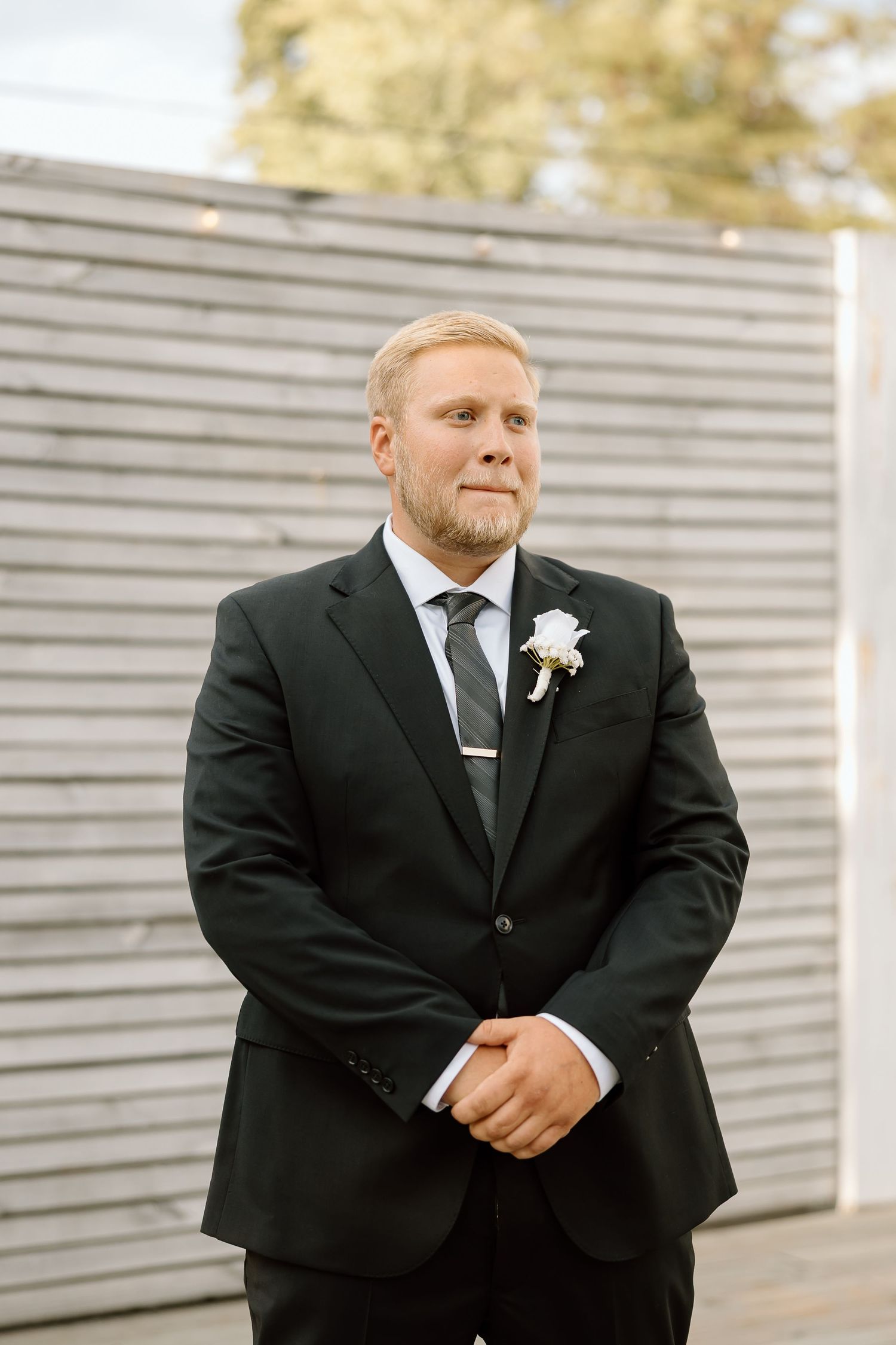 A person in a black suit with a white boutonniere poses against a light-colored wall.