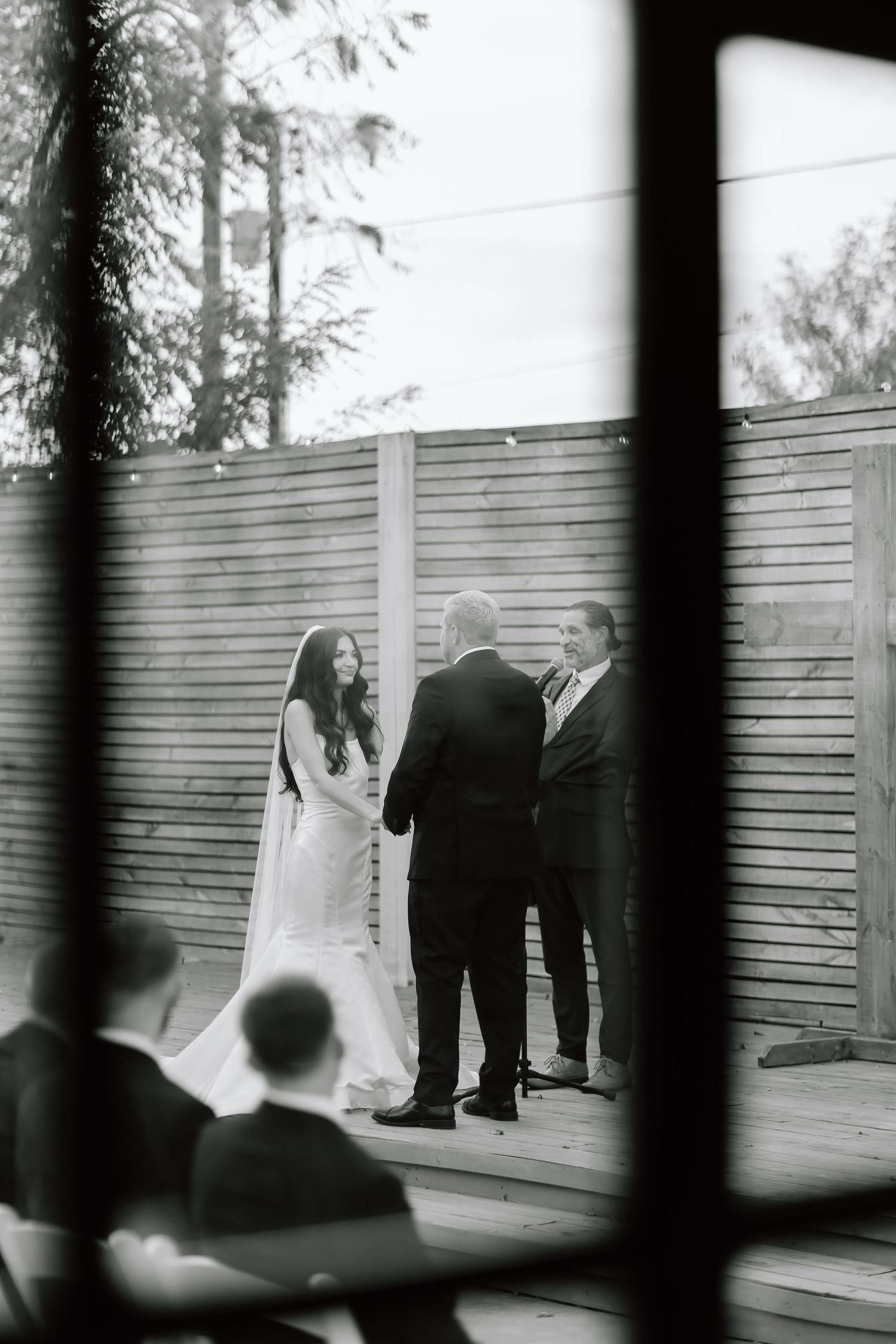 A black and white wedding photo showing guests gathered on a wooden deck framed through vertical railings.