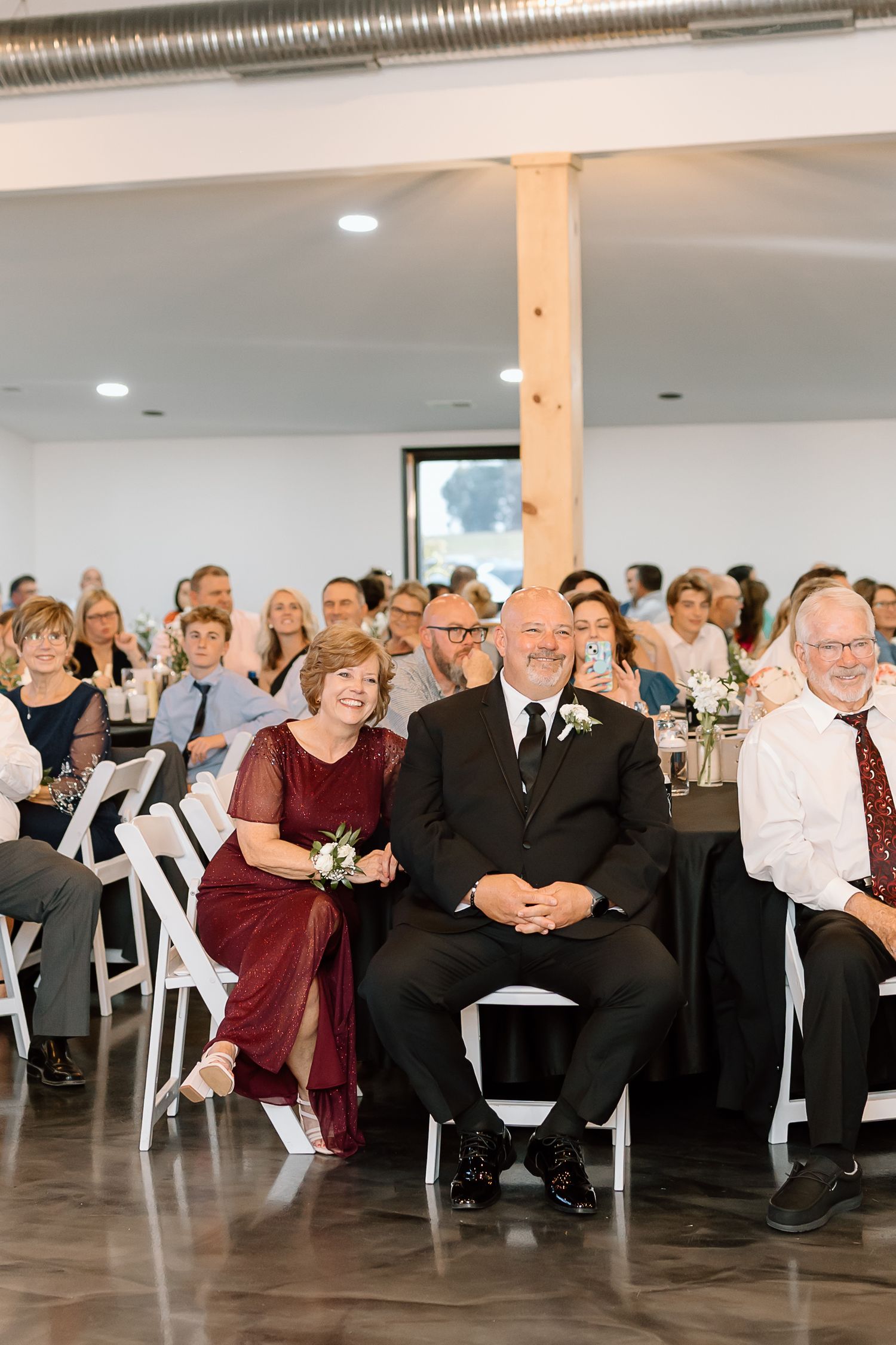Wedding guests seated in rows of white chairs watch attentively during an indoor ceremony.