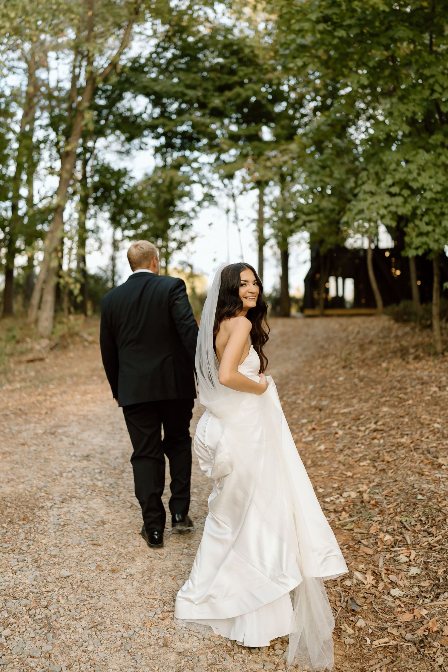 A bride and groom walk away together down a tree-lined path in a romantic woodland setting.