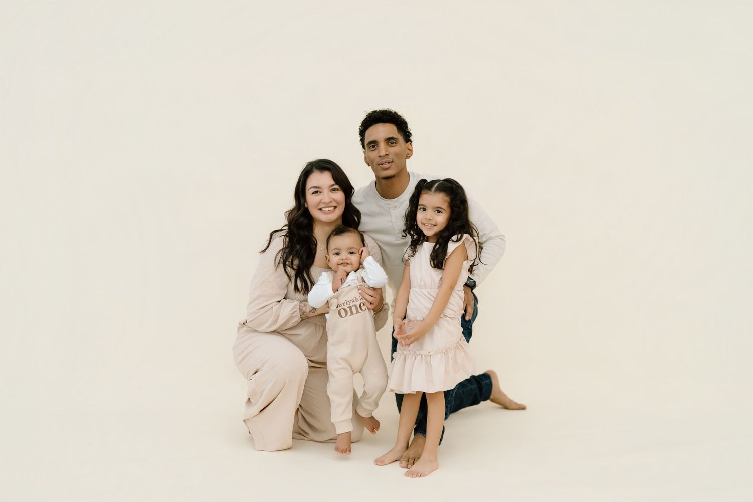A family in coordinated neutral outfits poses together against a cream colored backdrop for a studio portrait.