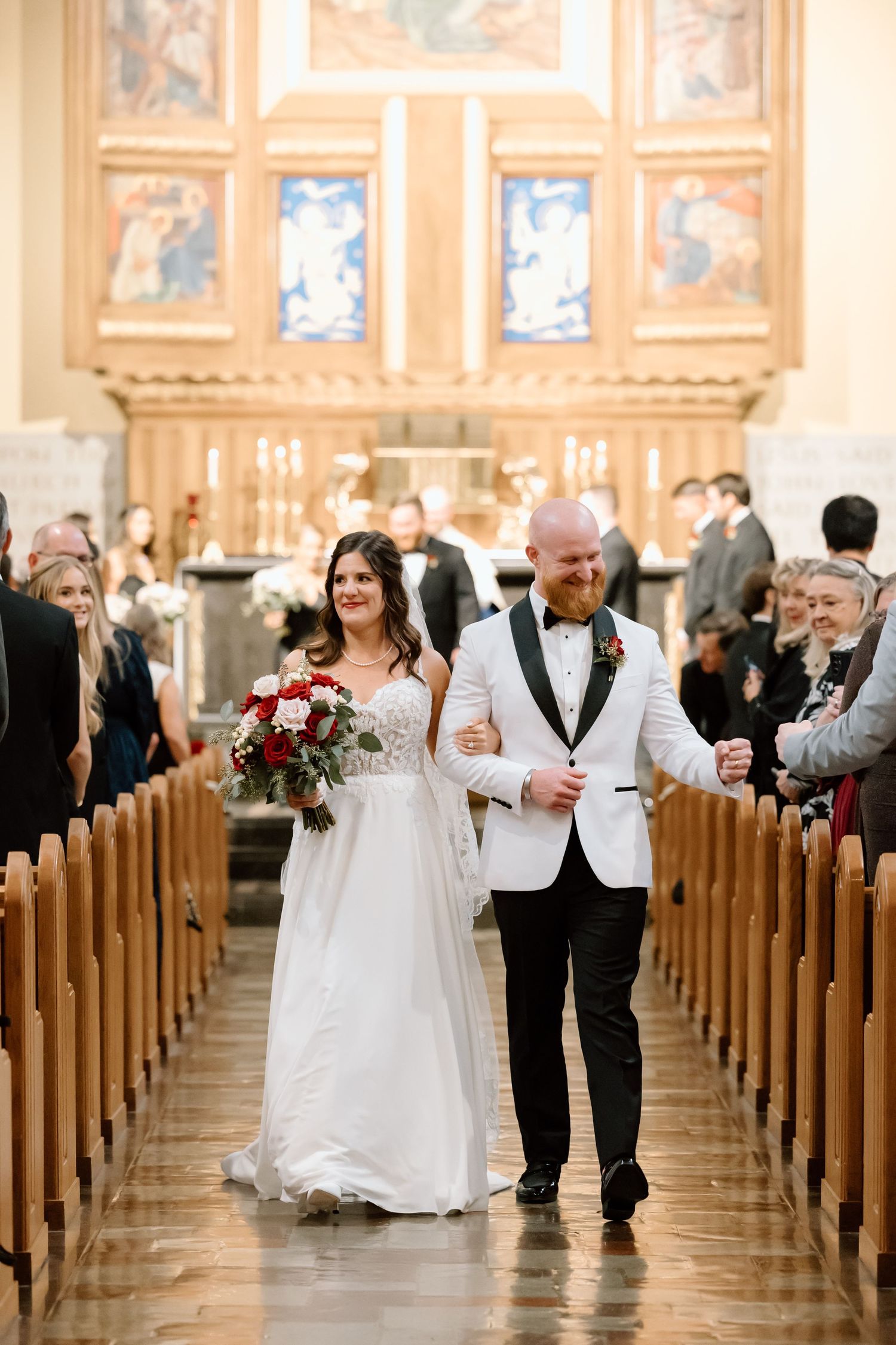 Newlyweds walk down the aisle of an ornate church with wooden pews and decorative altar after their wedding ceremony.