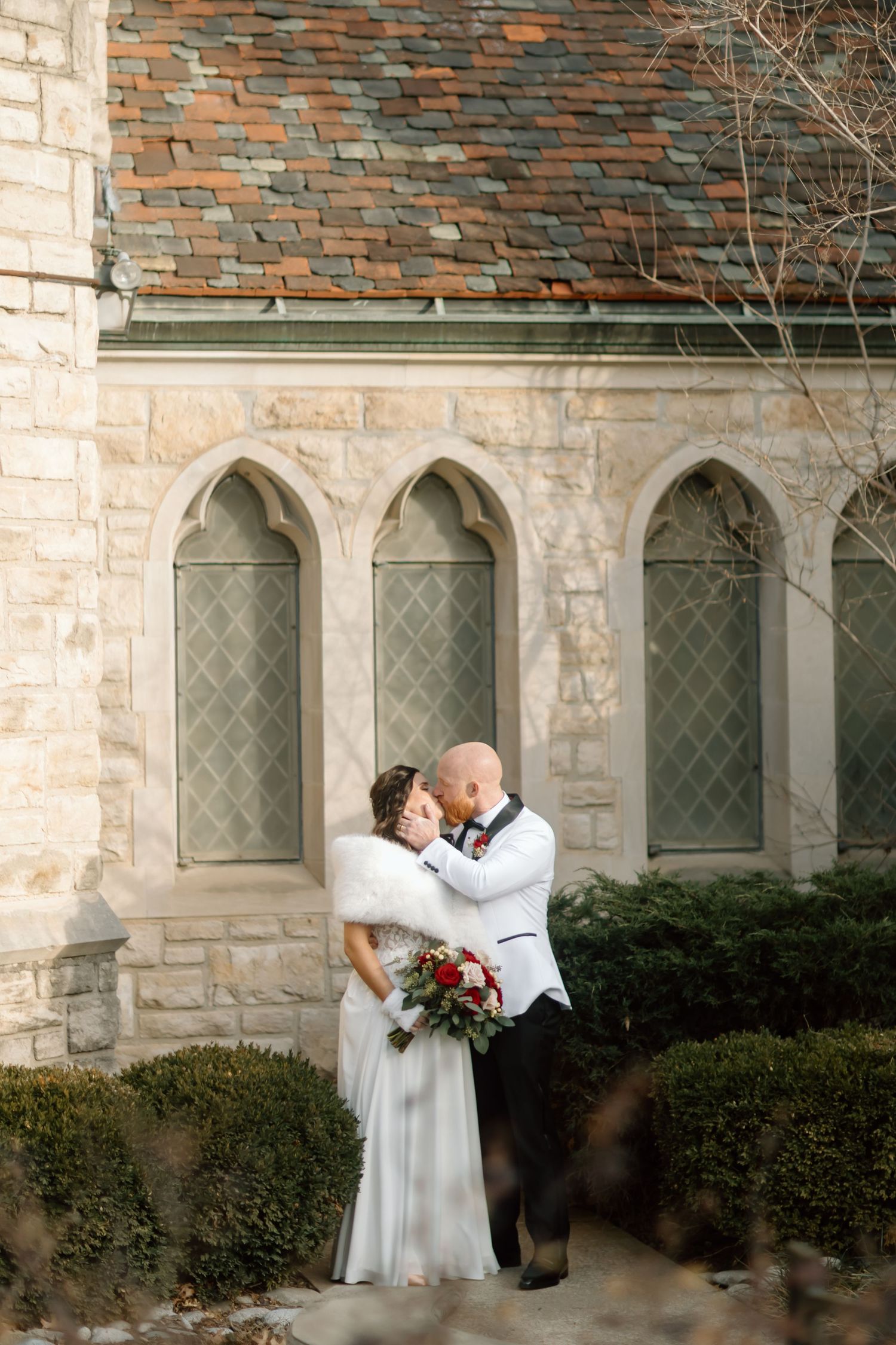A romantic wedding couple shares intimate moments against a stone church wall with arched windows.