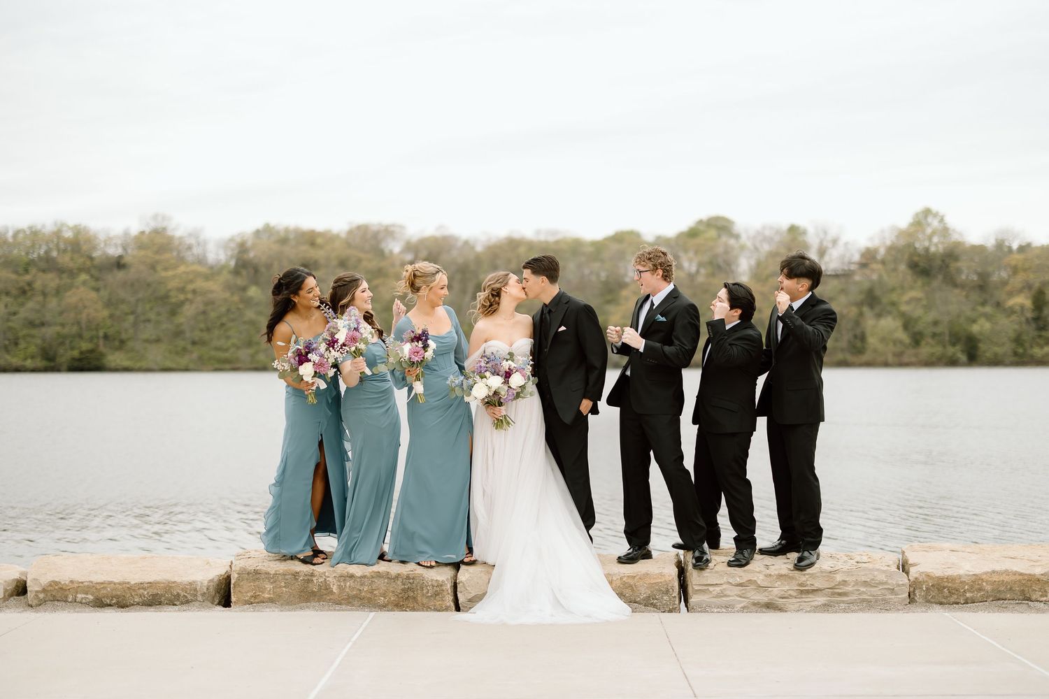 Wedding party in blue bridesmaid dresses and black suits pose by lakeside.