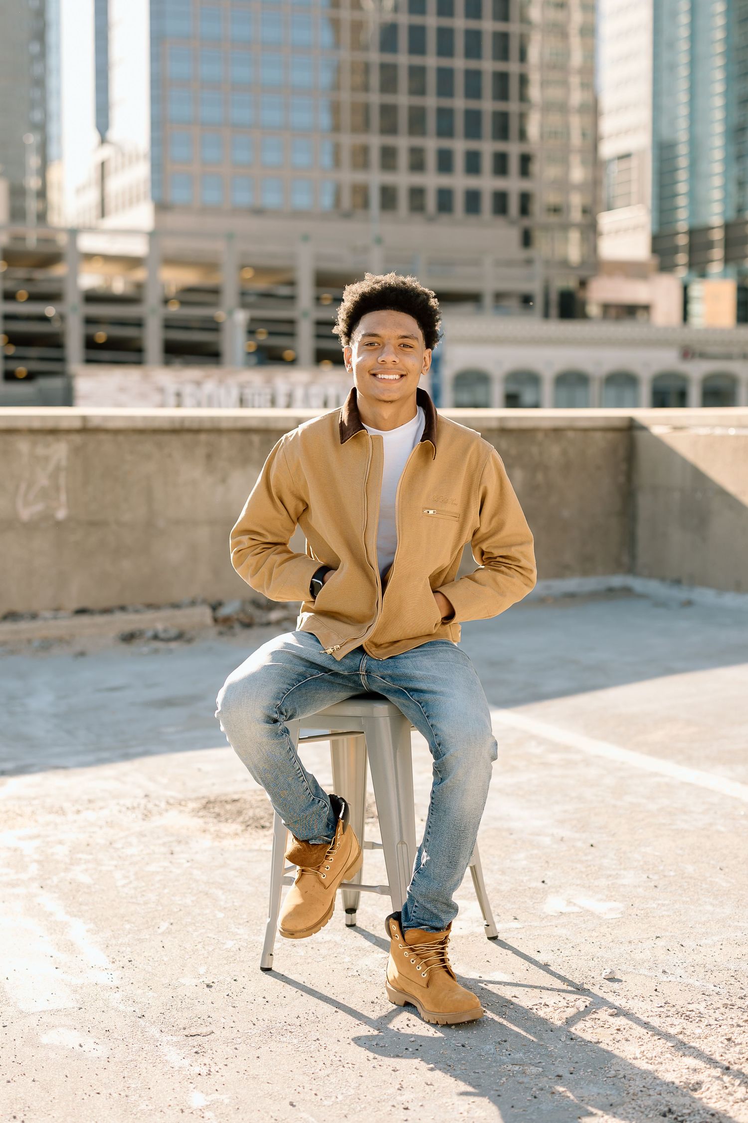 Person in casual wear sits on stool against city backdrop.