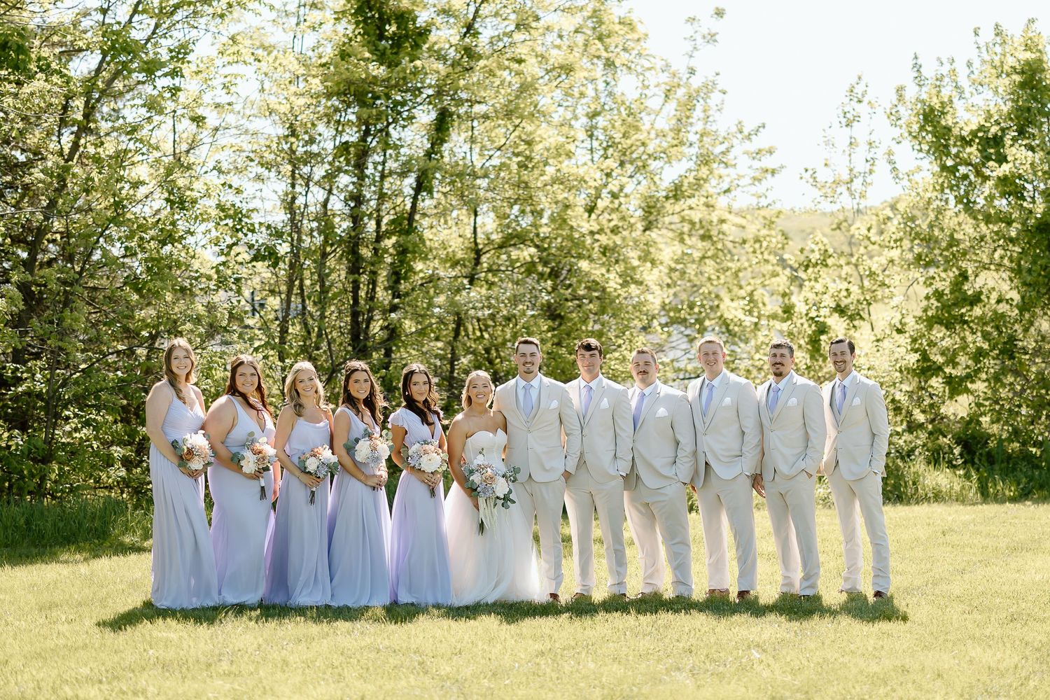 Wedding party in light blue dresses and beige suits poses on grass with trees in background.