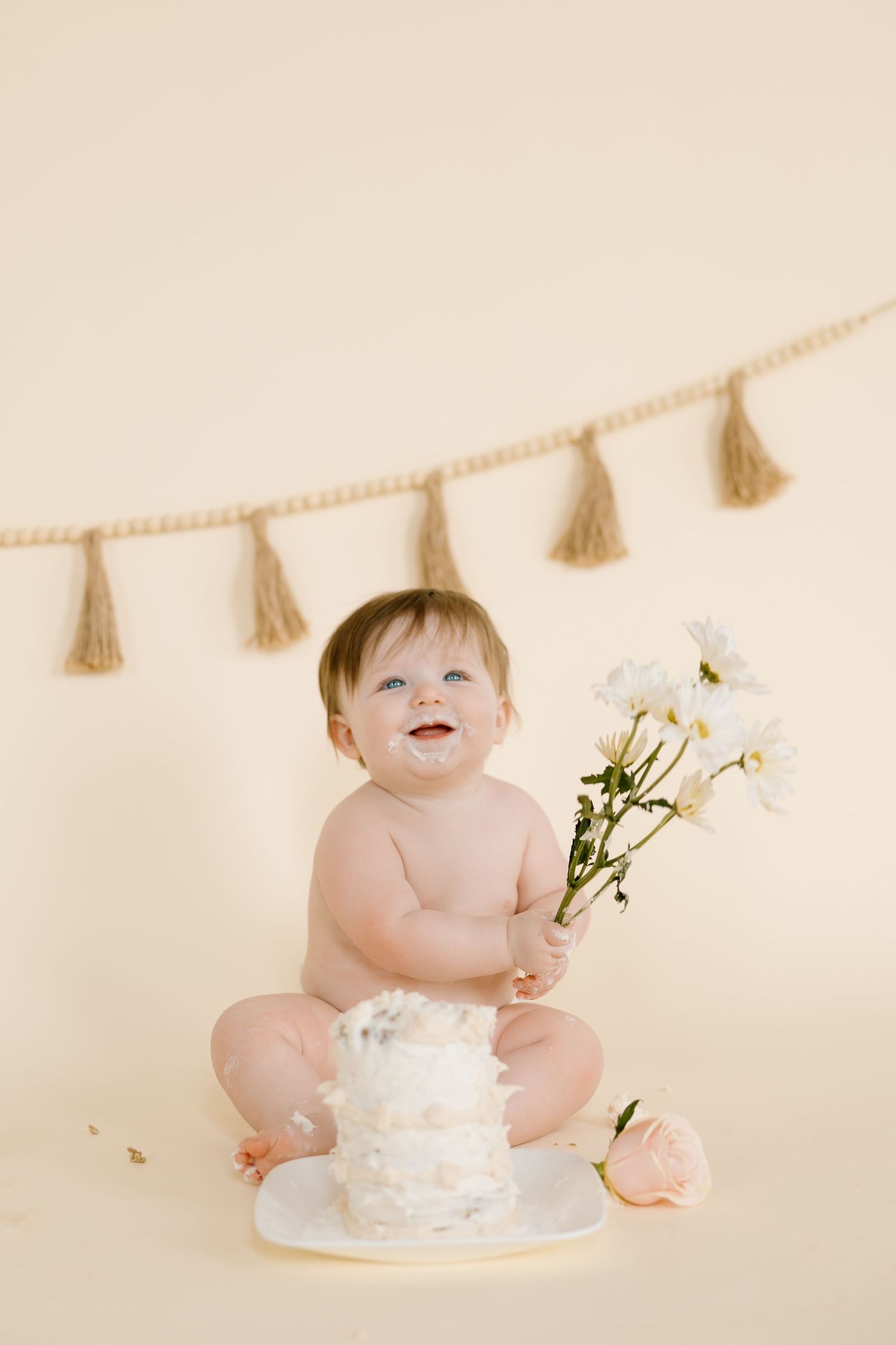 Baby sits with white flowers and cake during cake smash photoshoot with tassel banner decoration.