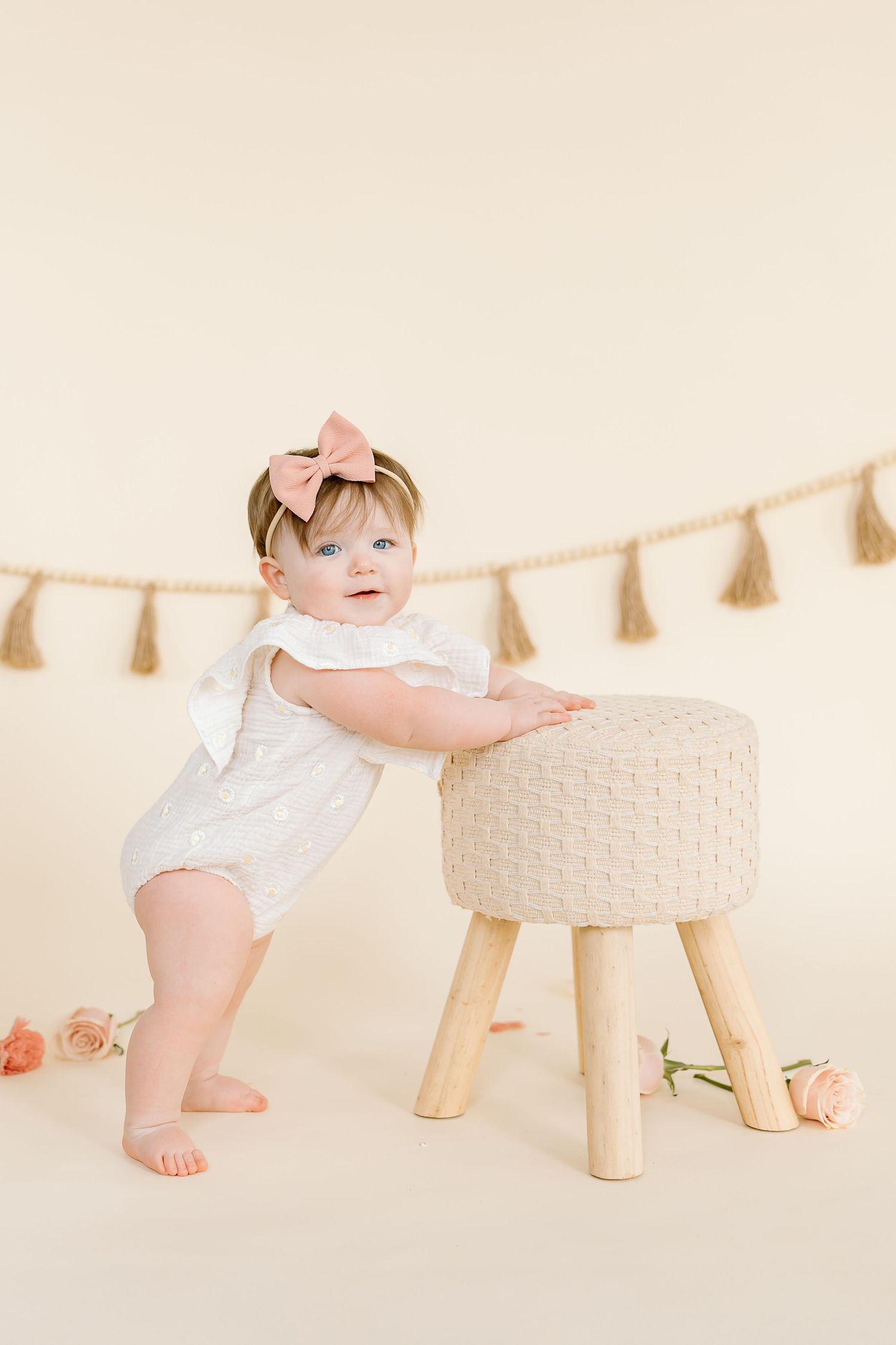 Small child in white romper leans on knitted stool beneath decorative tassels.