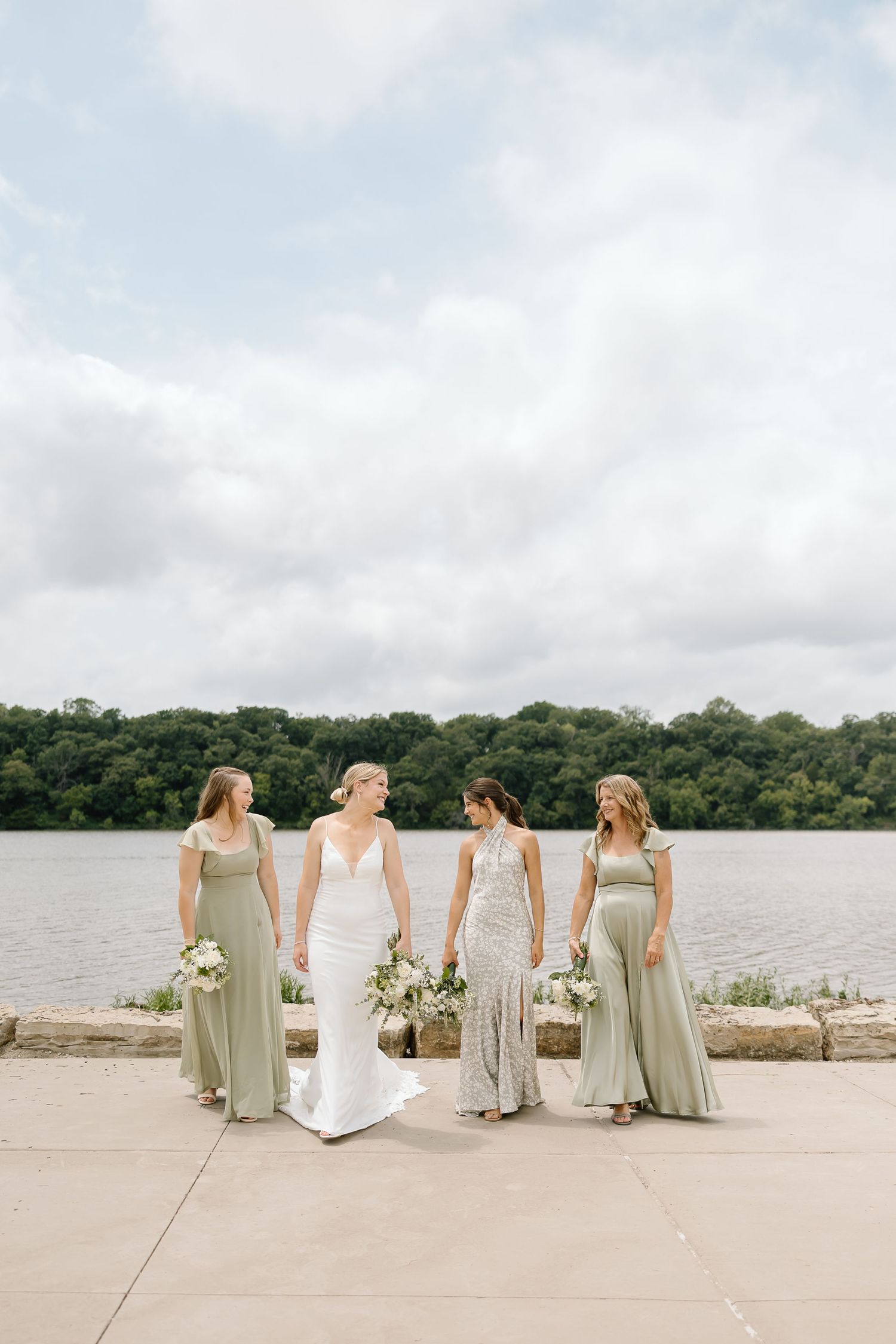 Wedding party wearing sage green and white dresses poses together on concrete patio by a lake under cloudy skies.