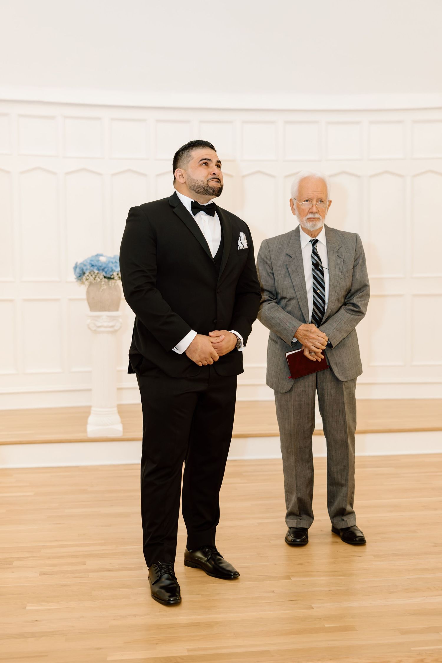 Two men in formal attire stand in an elegant white wedding venue, one in a tuxedo and one in a gray suit.