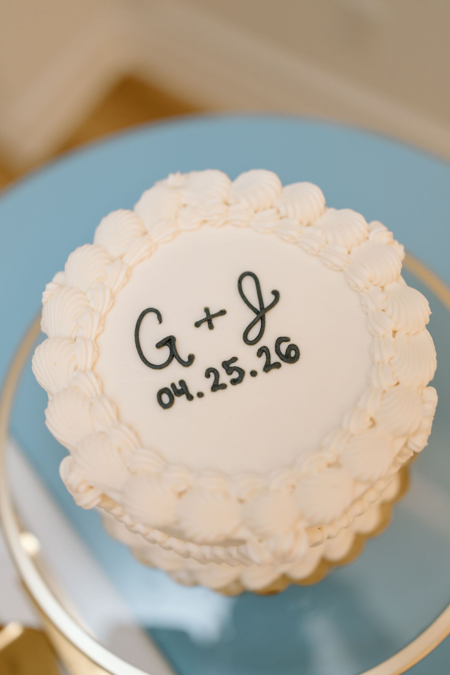 White wedding cake with scalloped frosting, monogram G+J and date 04.25.26 on blue cake stand.