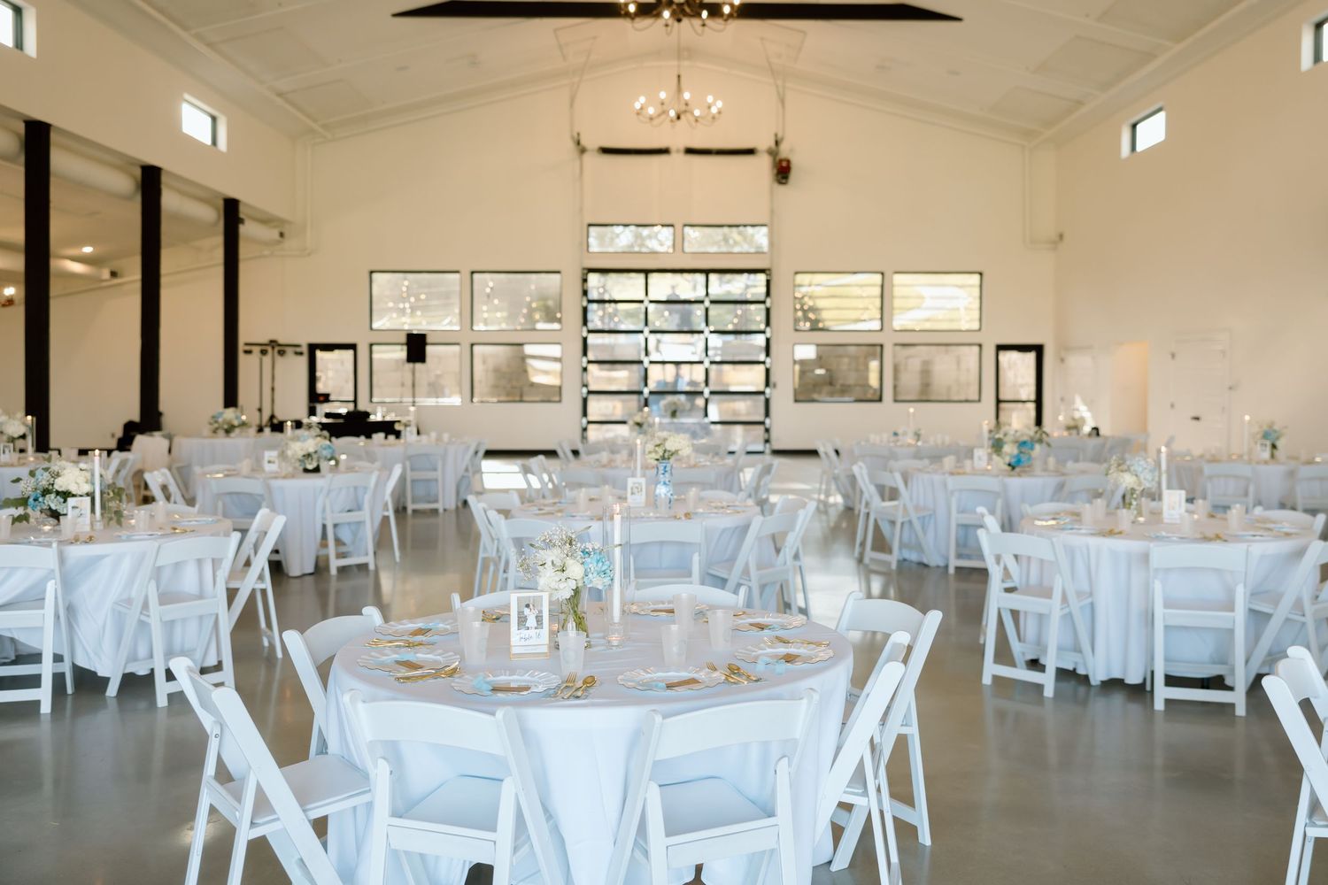 Elegant wedding reception hall with white round tables, chairs, and chandelier lighting in a bright, airy venue.