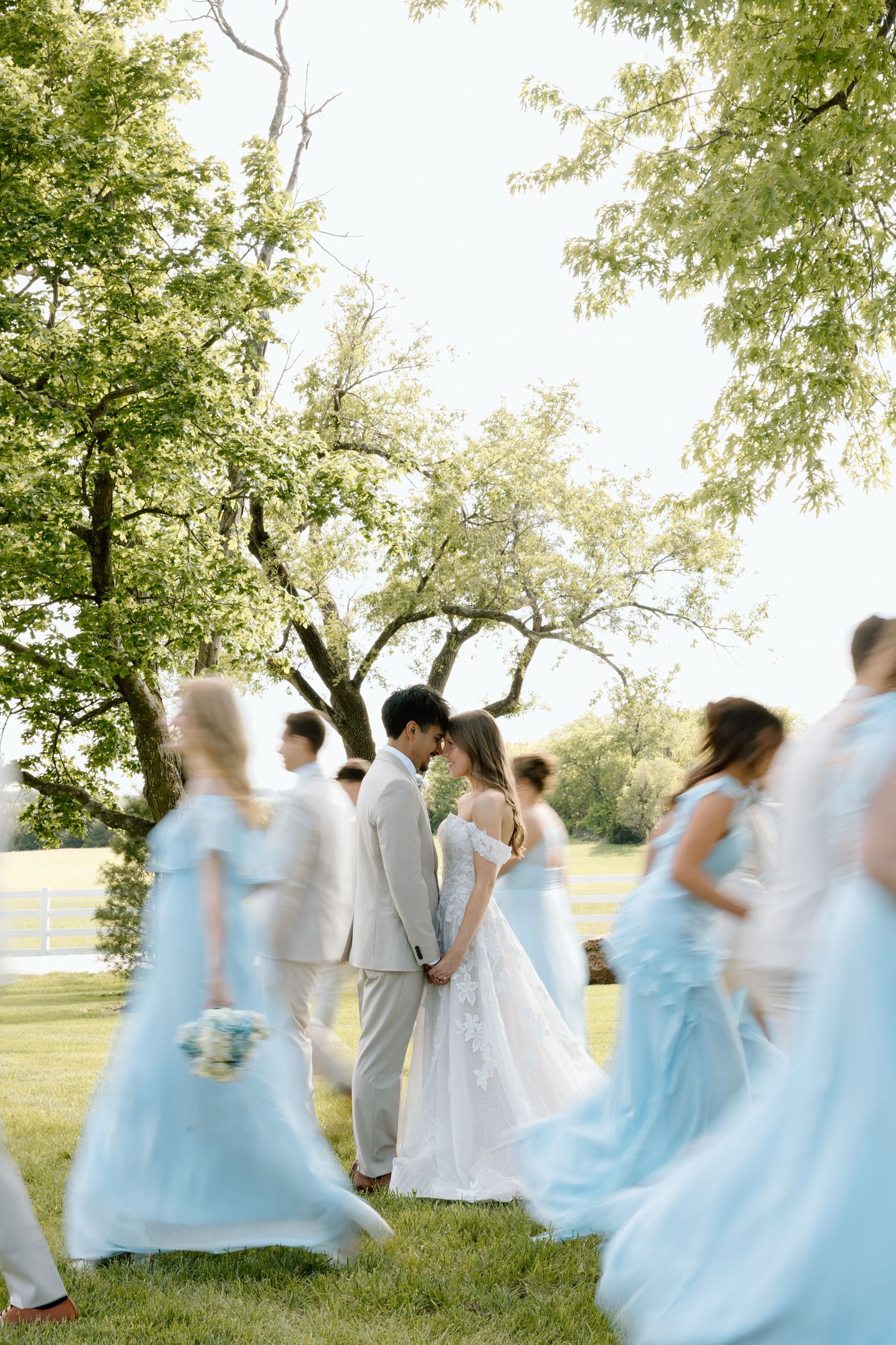 Bride and groom share an intimate moment surrounded by blurred wedding party in light blue dresses outdoors.