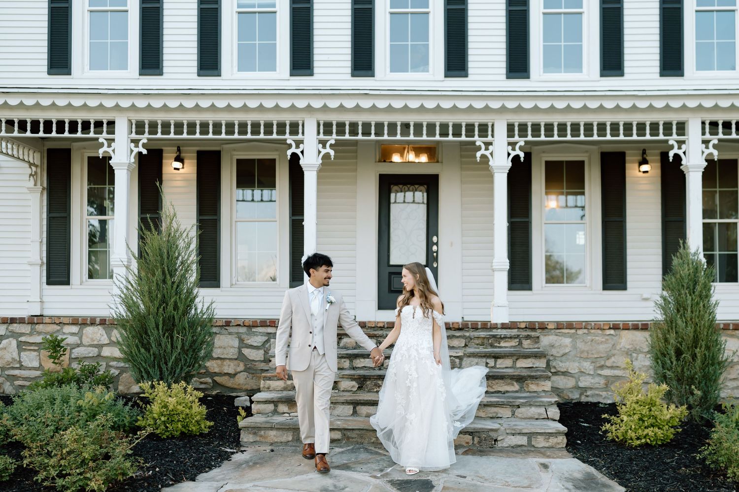 Bride and groom hold hands on stone steps before a white Victorian farmhouse with dark shutters and porch columns.