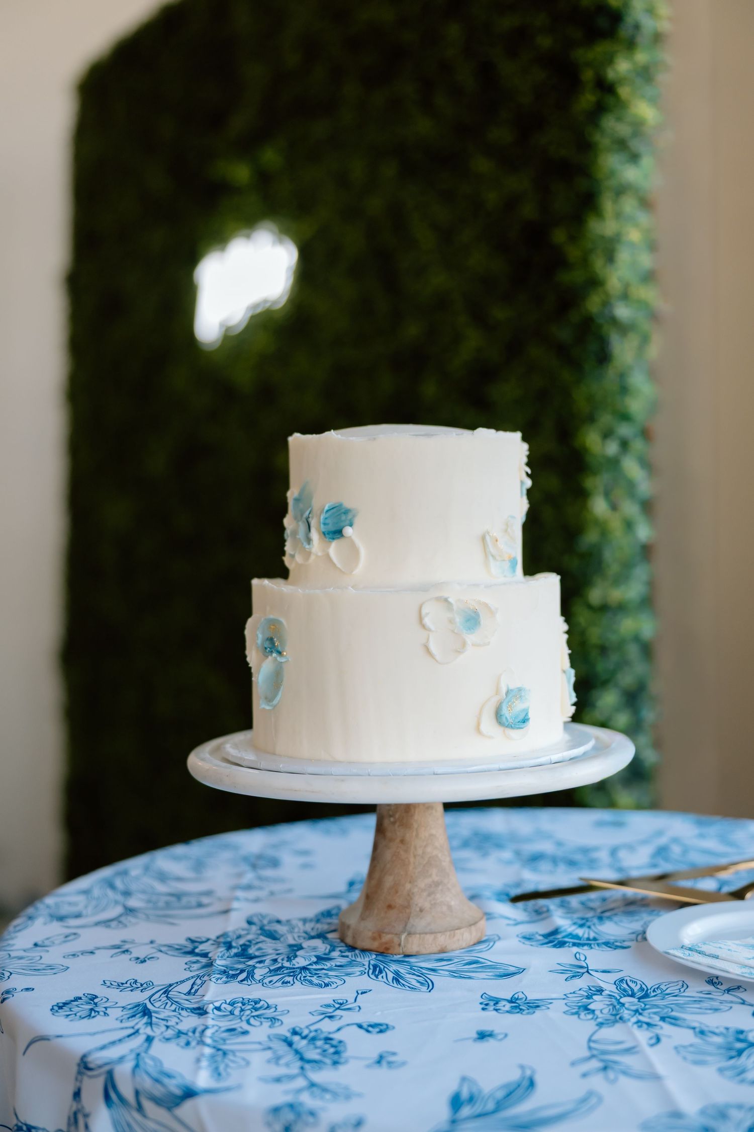 Two-tier white wedding cake with blue floral accents on a wooden stand, set against a green hedge backdrop.