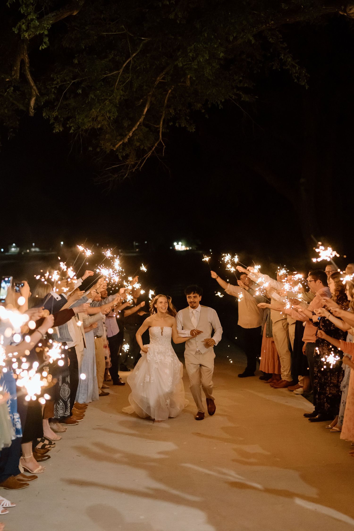 Bride and groom exit through sparkler tunnel surrounded by wedding guests at night outdoor ceremony.