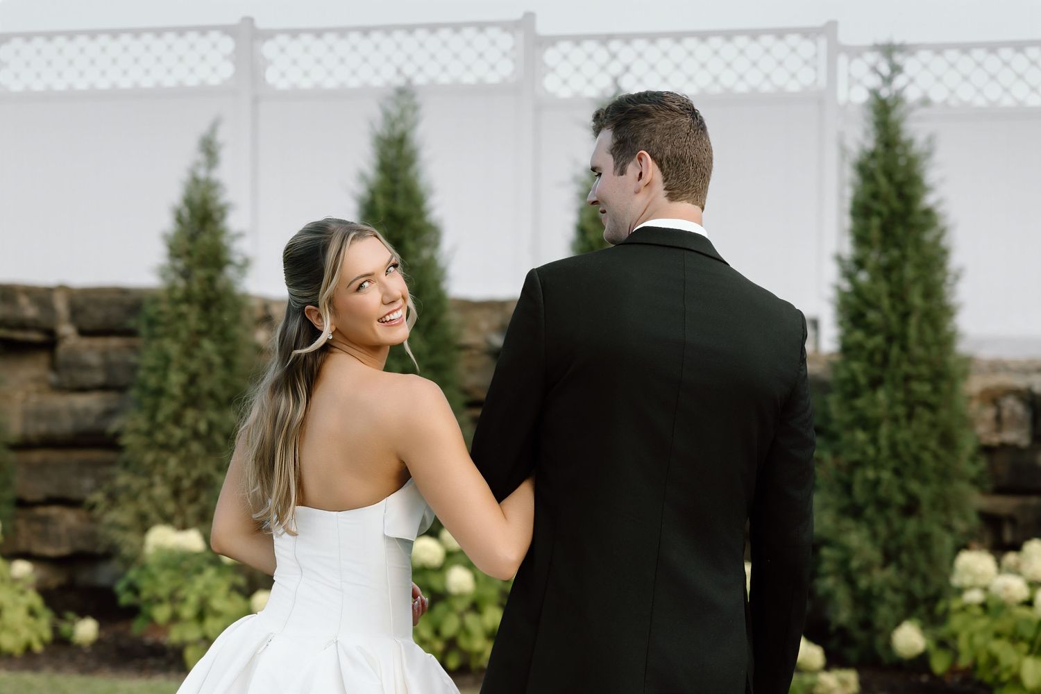 Bride in white strapless gown smiles over shoulder at groom in black tuxedo, holding hands in garden setting.