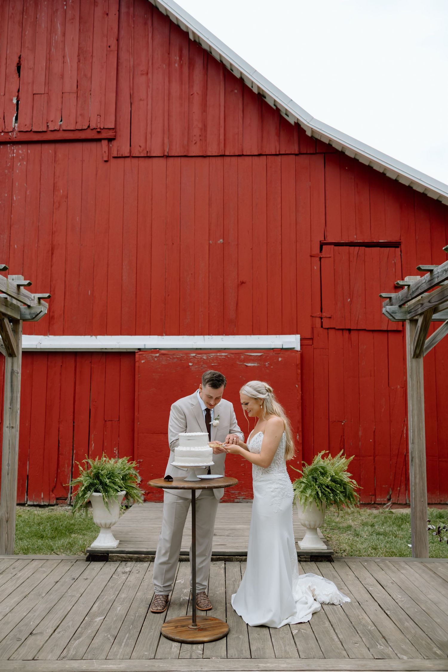 Bride and groom cutting wedding cake on rustic wooden deck in front of a classic red barn.