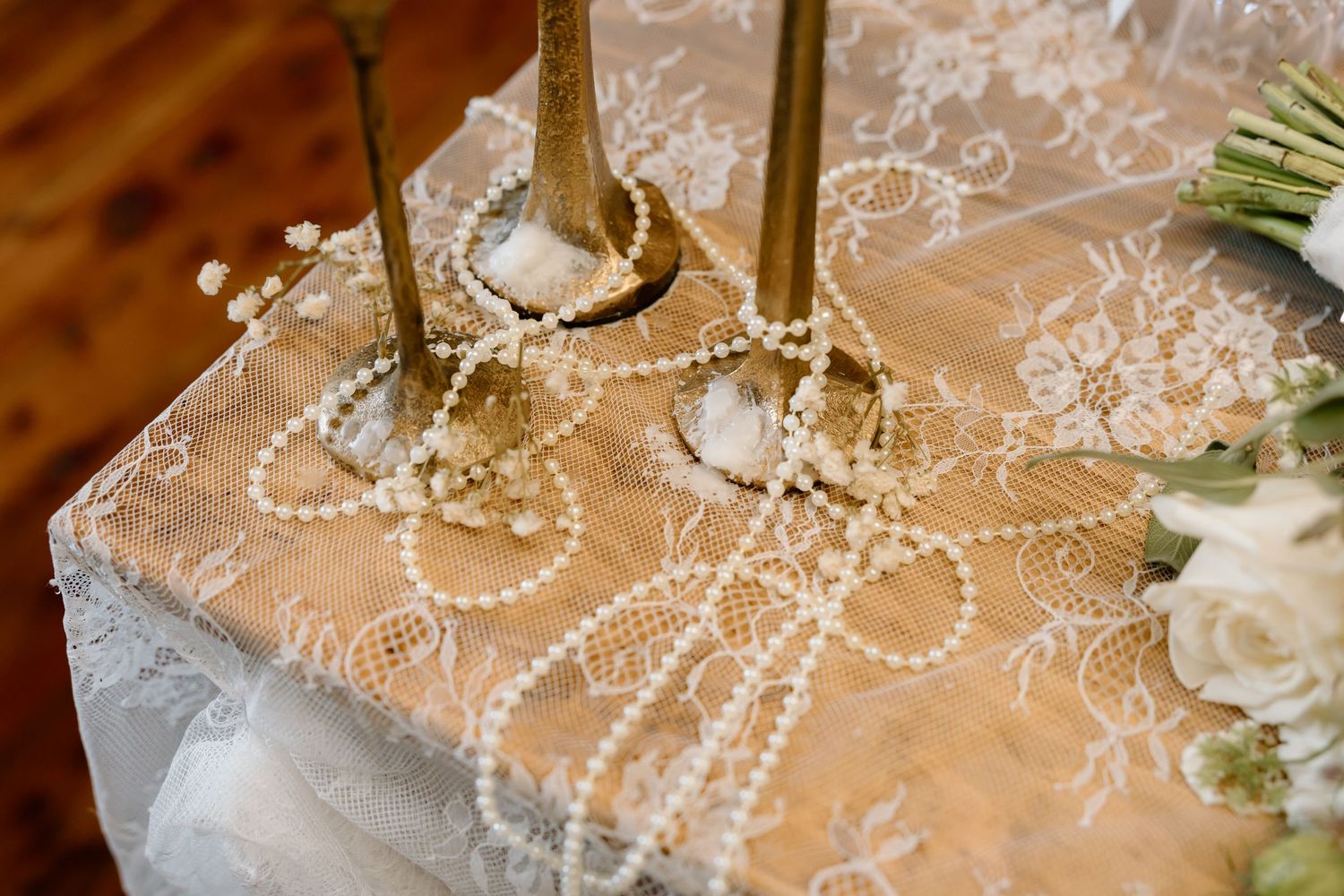 Elegant wedding table with lace overlay, pearl necklace, gold candlesticks, and white floral bouquet.