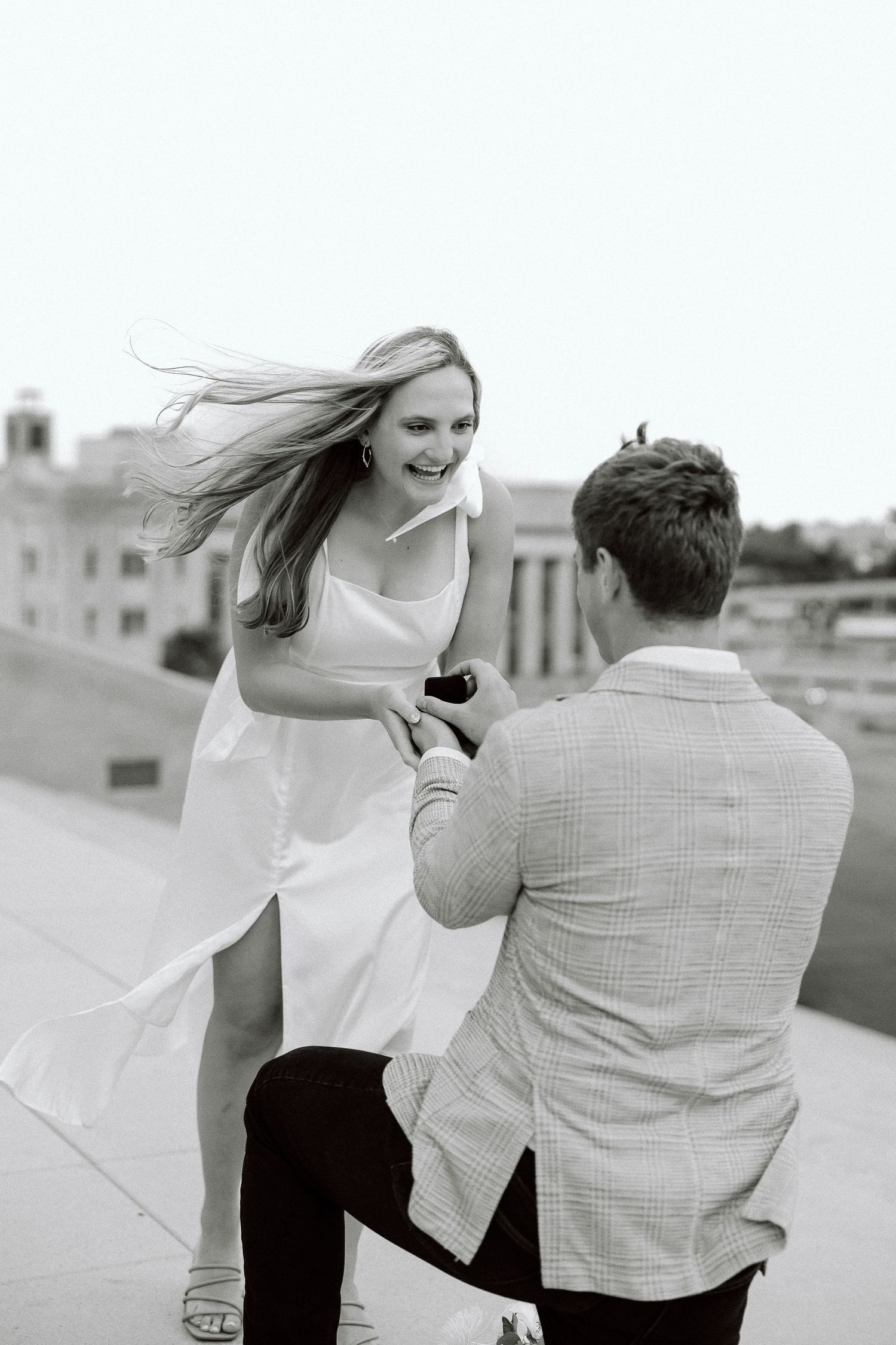 A couple in black and white dance together on a rooftop, her white dress flowing in the breeze.