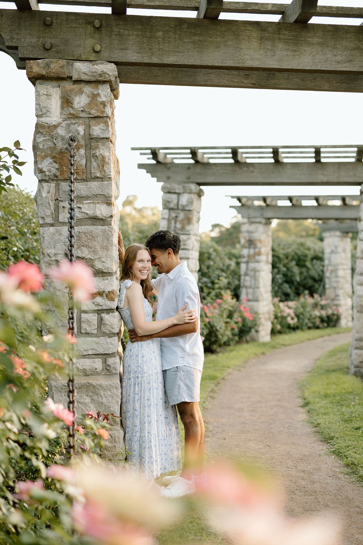 A romantic couple shares tender moments under a pergola surrounded by blooming flowers in a garden setting.