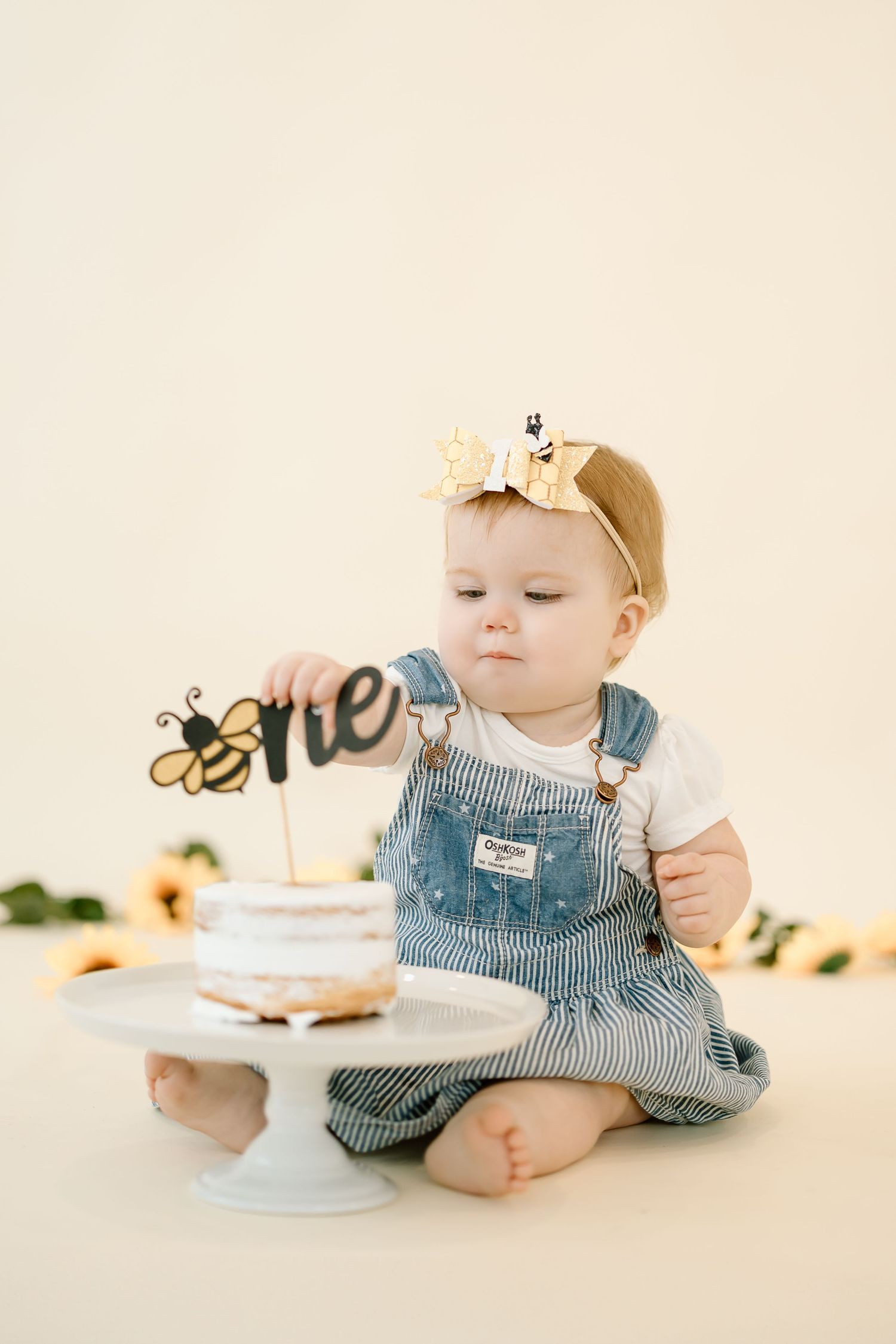 Baby in blue overalls sits beside first birthday cake with bee-themed topper on white cake stand.