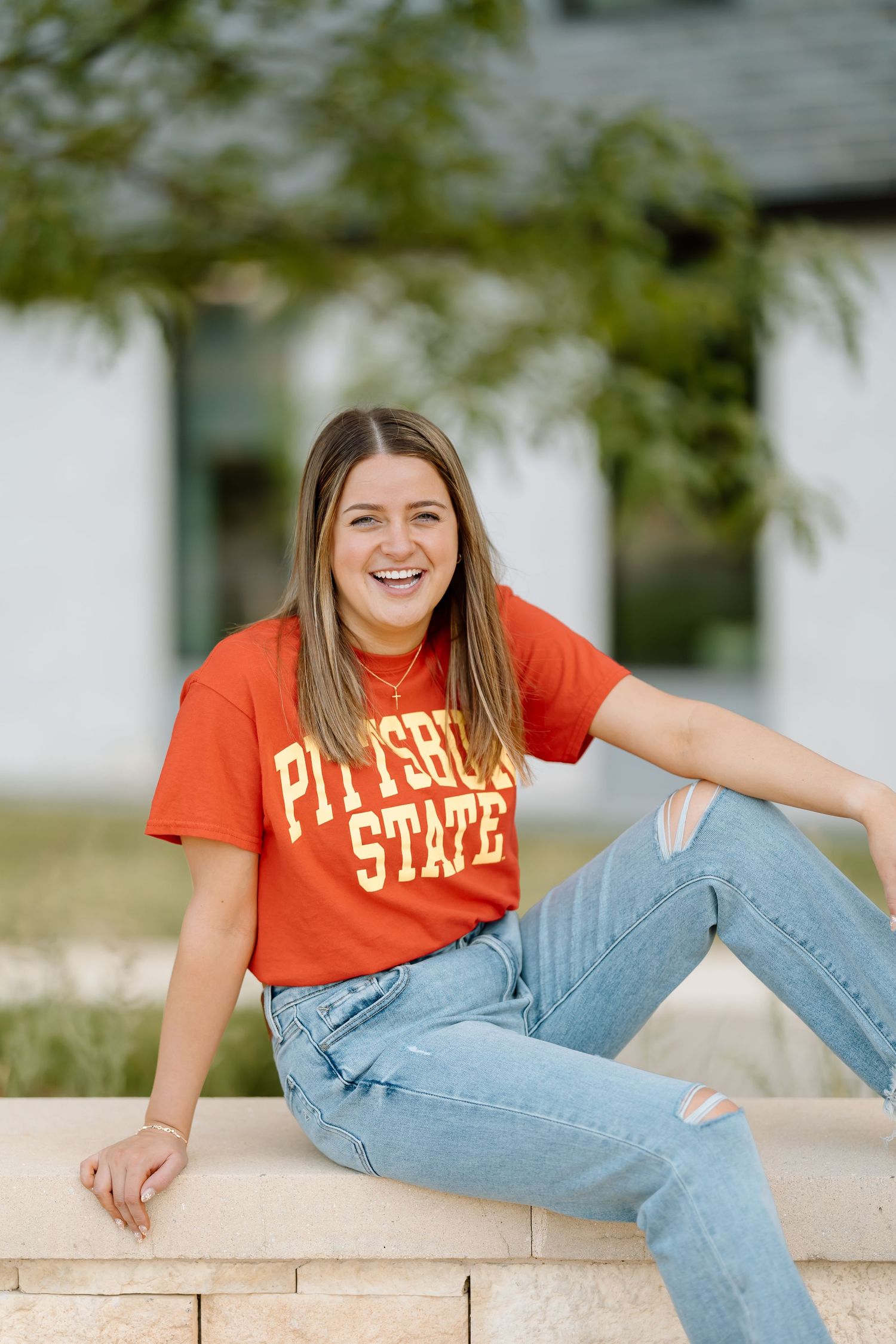 Student wearing orange Pittsburg State t-shirt and jeans sits casually on stone wall outdoors on sunny day.